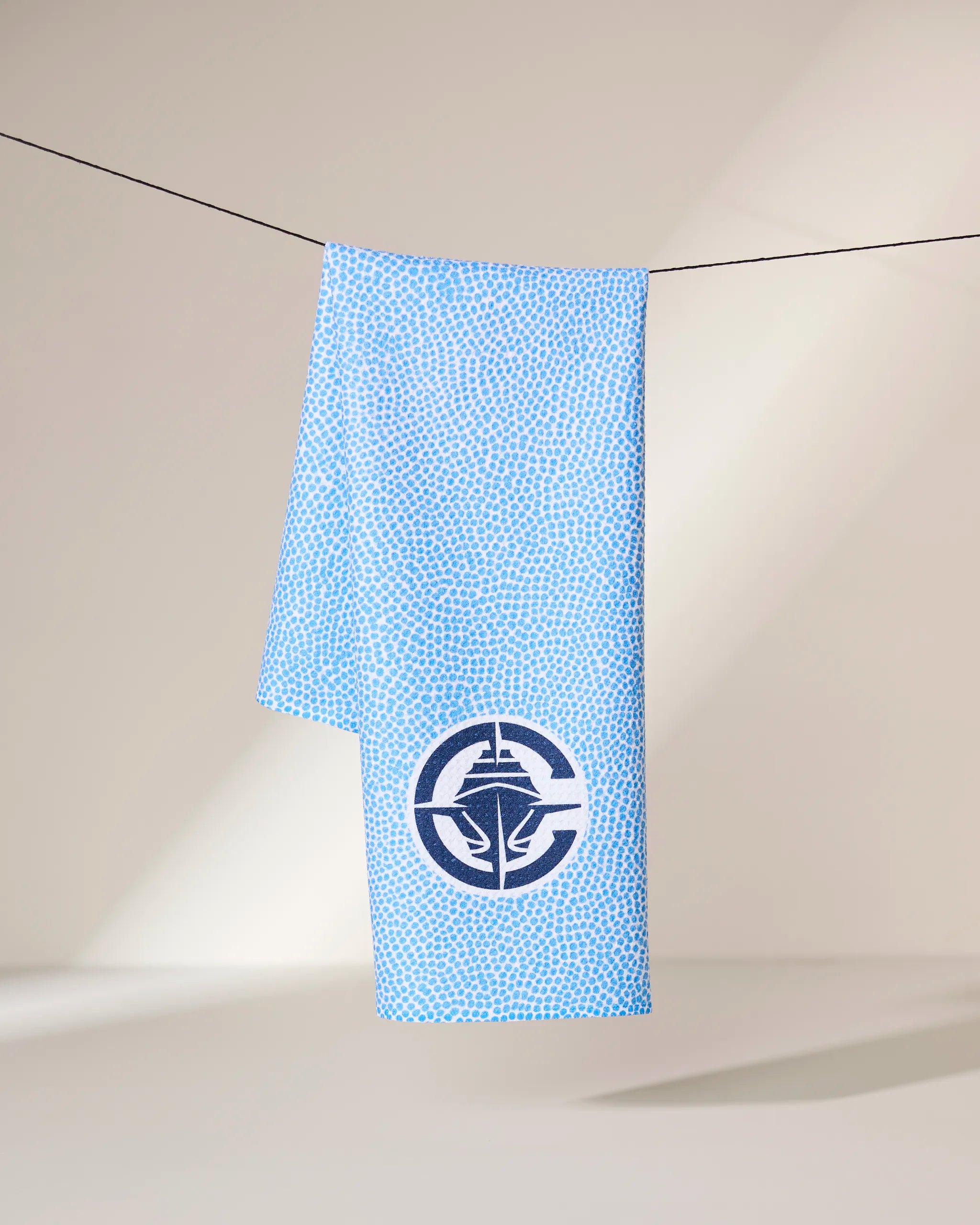 A blue Clippers Grip towel by Geometry, featuring a subtle dotted pattern and a circular logo with a stylized leaf, hangs on a black clothesline against a neutral background.