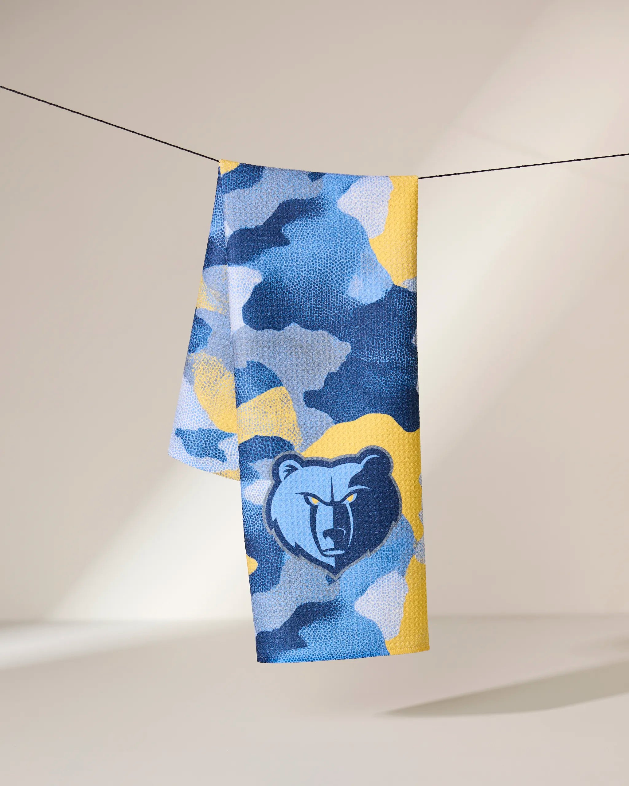 The Geometry Grizzlies Camo towel, featuring a blue, yellow, and gray camouflage design with the Memphis Grizzlies bear logo, hangs over a black line against a neutral background.