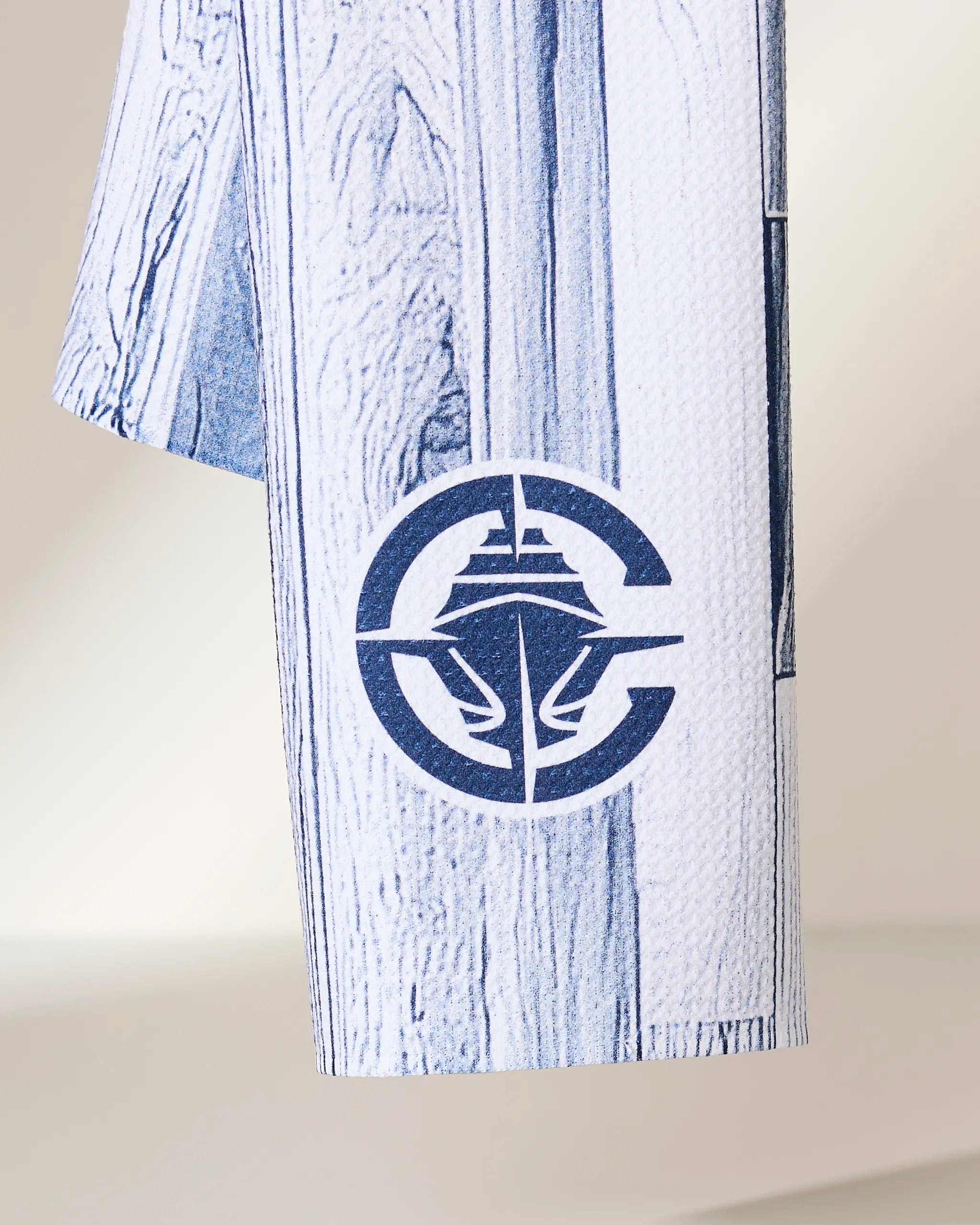 A white and blue textured fabric from Geometry, called Clippers Zone, featuring a printed circular logo with a stylized ship and waves, hanging against a neutral background.