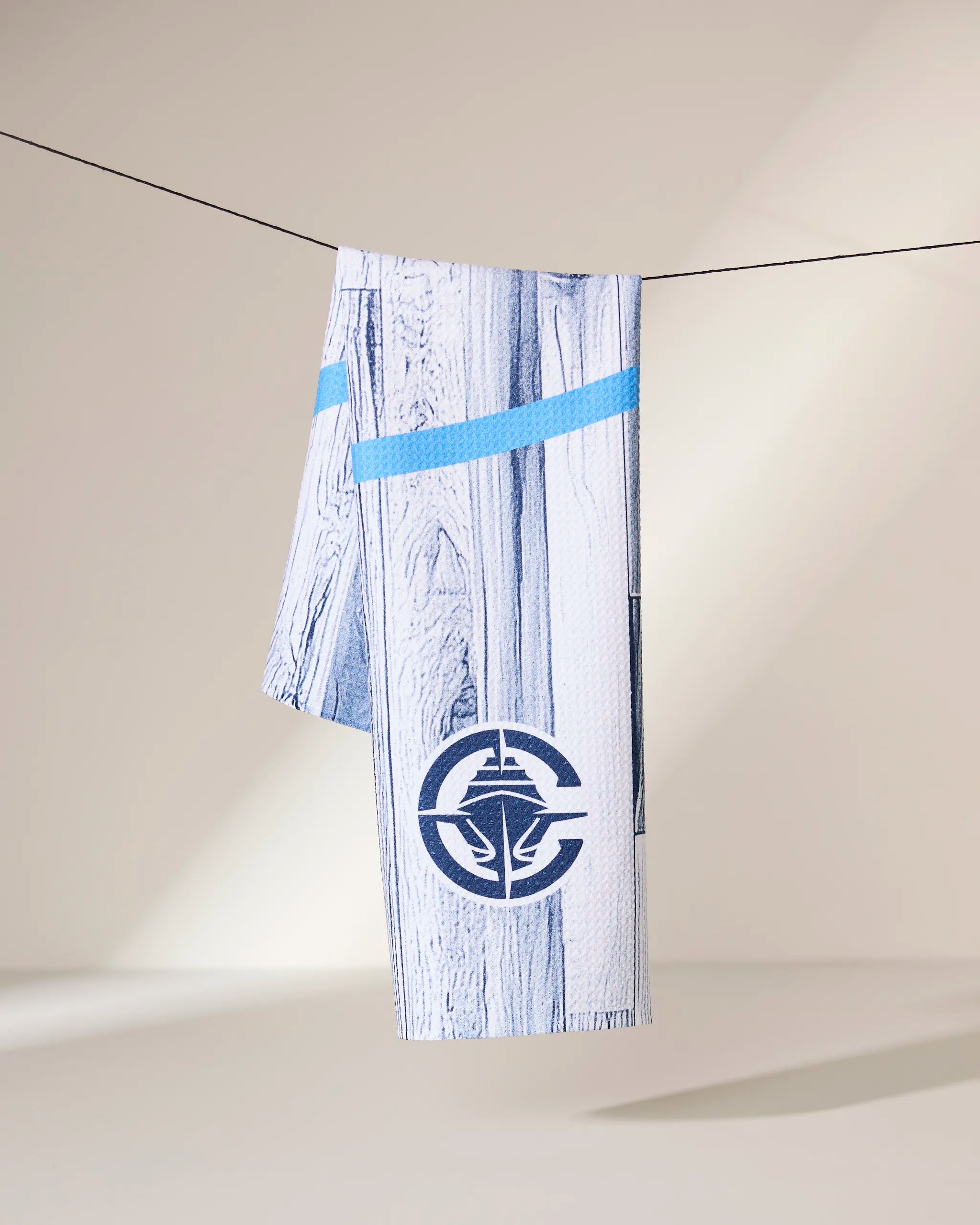 A Geometry Clippers Zone towel, white with a blue stripe and a round blue fish logo, hangs over a black clothesline against a neutral background.