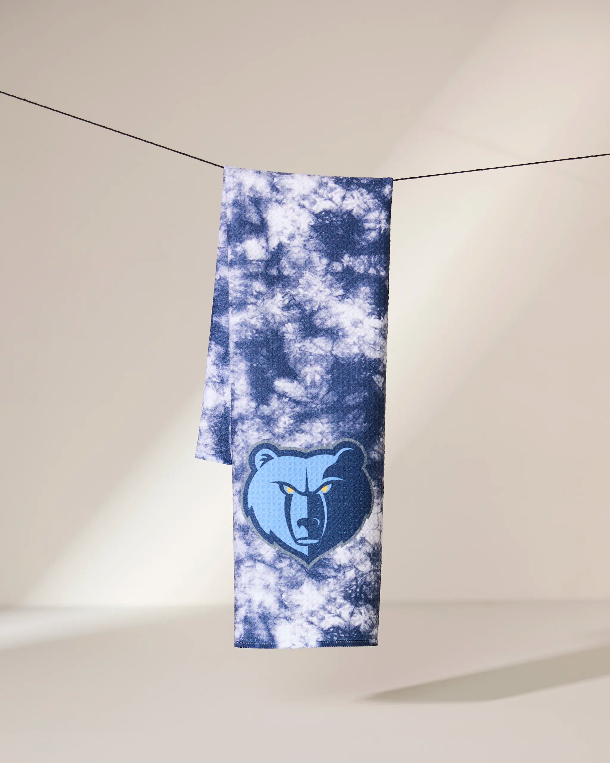 The Grizzlies Flash towel by Geometry, showcasing a blue and white tie-dye design with the Memphis Grizzlies bear logo, hangs on a black clothesline against a neutral background.