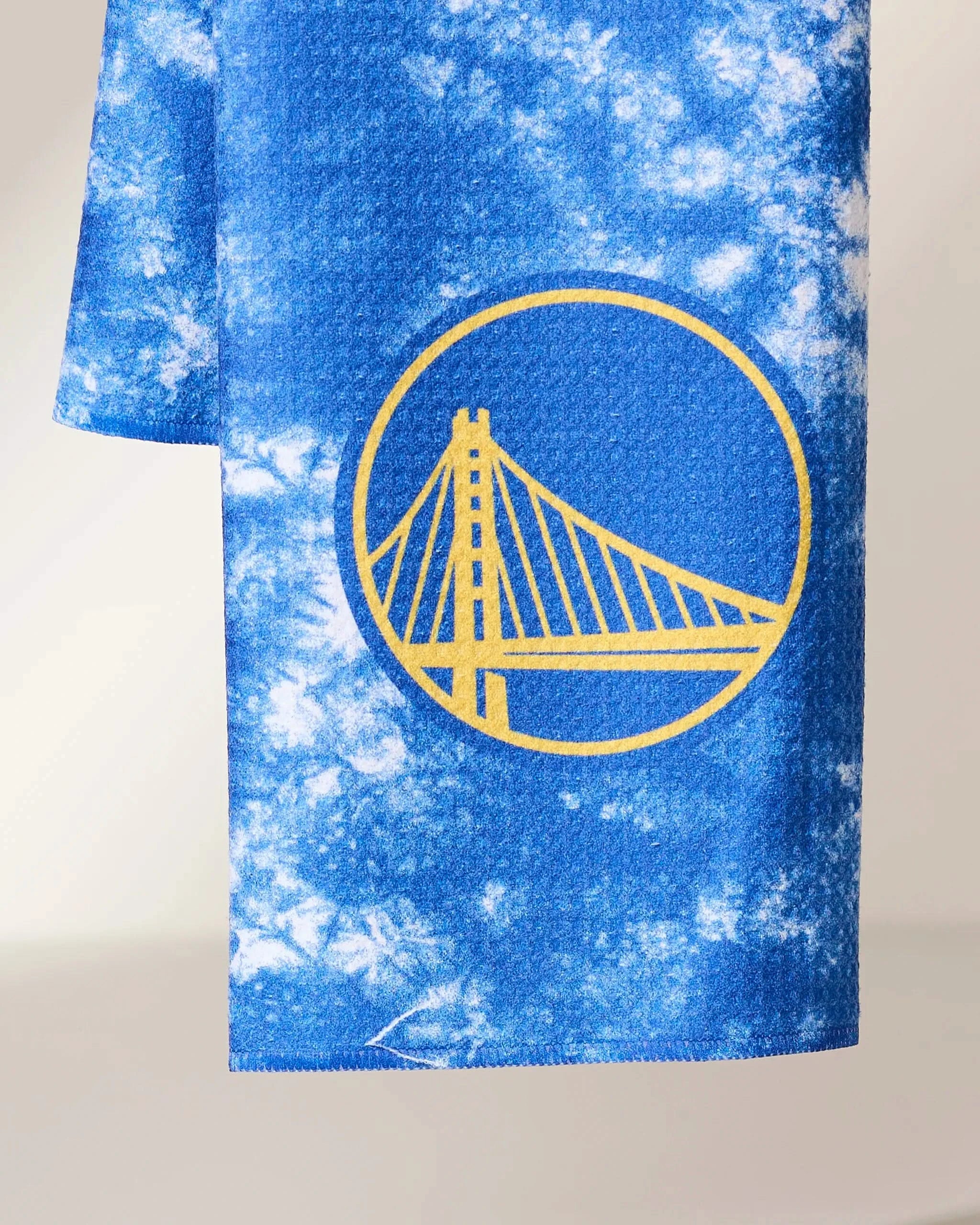 The Geometry Warriors Flash towel, in blue and white tie-dye with a yellow Golden Gate Bridge and Golden State Warriors logo, hangs against a neutral background.