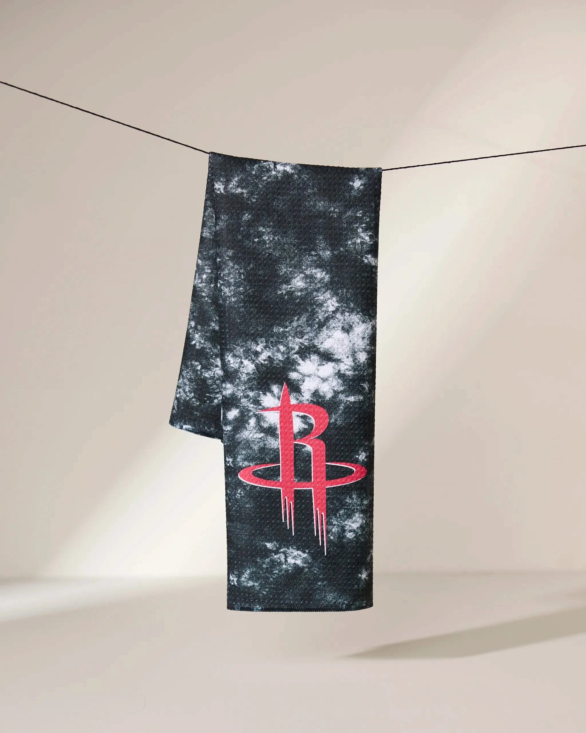 The Geometry Rockets Flash towel, in black and white tie-dye, hangs on a line with the red Houston Rockets logo near the bottom. The softly lit background is plain.