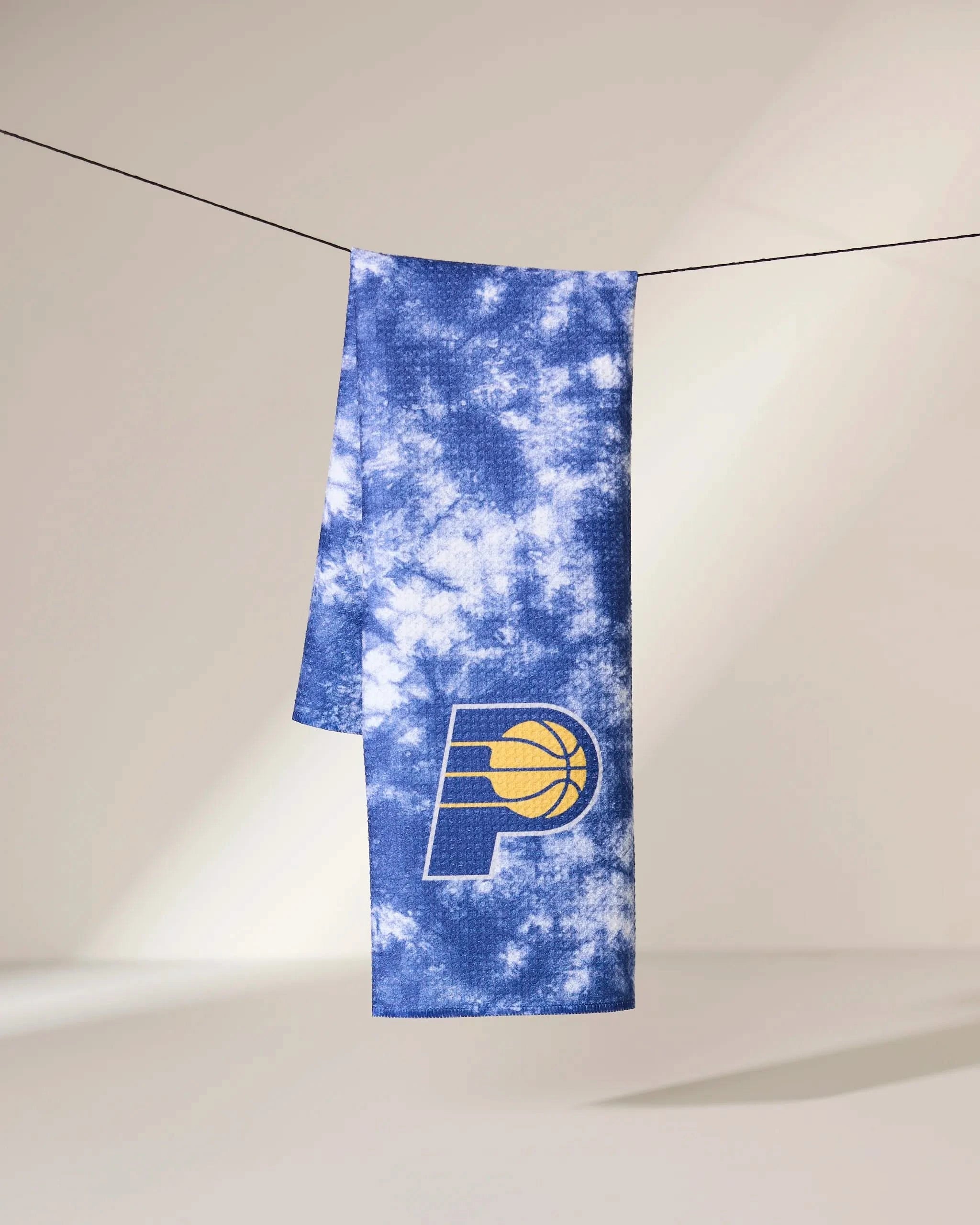 The Geometry Pacers Flash scarf, featuring a blue and white tie-dye design with the Indiana Pacers logo, hangs on a black string against a neutral background.