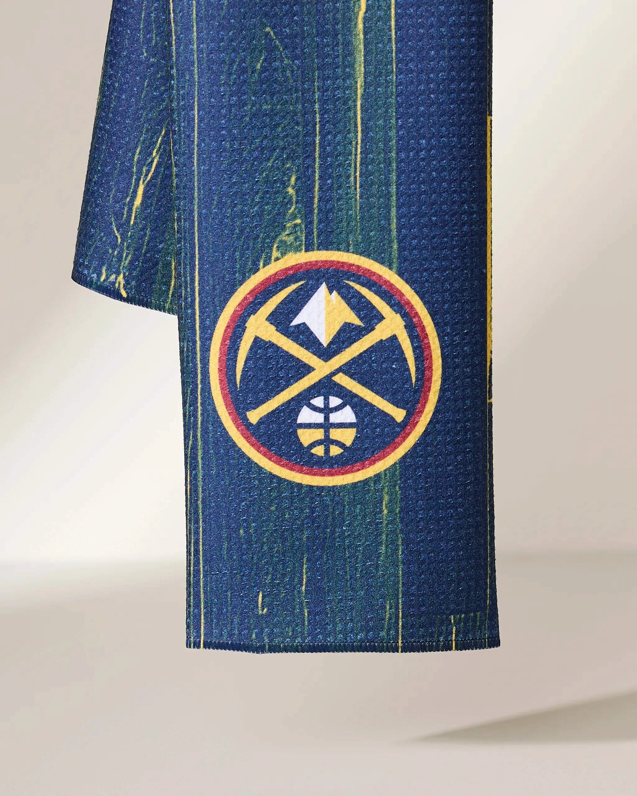 A close-up of the Geometry Nuggets Zone athletic towel, blue with subtle green and yellow streaks and a Denver Nuggets logo featuring crossed pickaxes and a mountain in yellow and red.