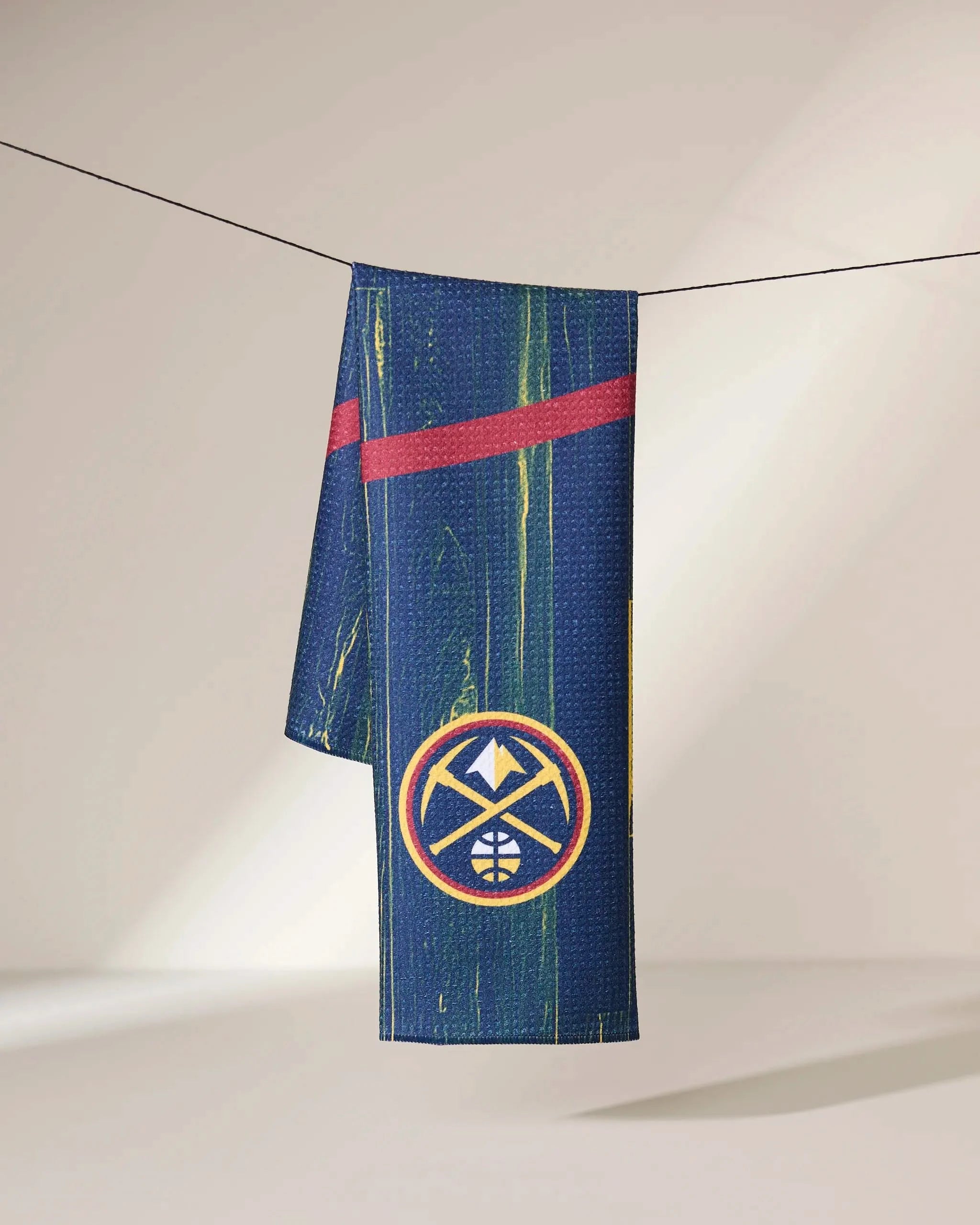 A blue Geometry Nuggets Zone towel, featuring the Denver Nuggets yellow and white logo with a red diagonal stripe, hangs on a clothesline against a neutral background.