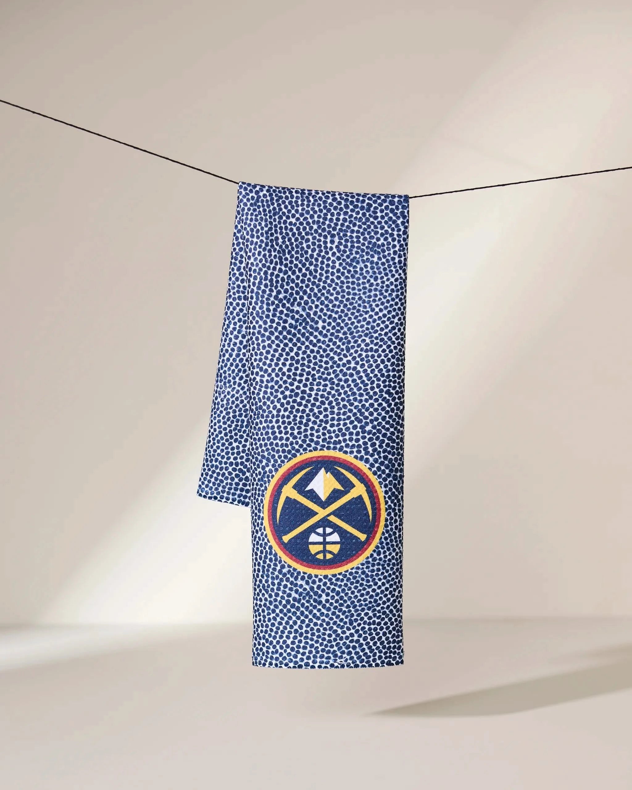 A blue and white speckled Geometry Nuggets Grip scarf hangs on a string, showcasing a circular patch with a yellow, red, and navy mountain-and-basketball logo.
