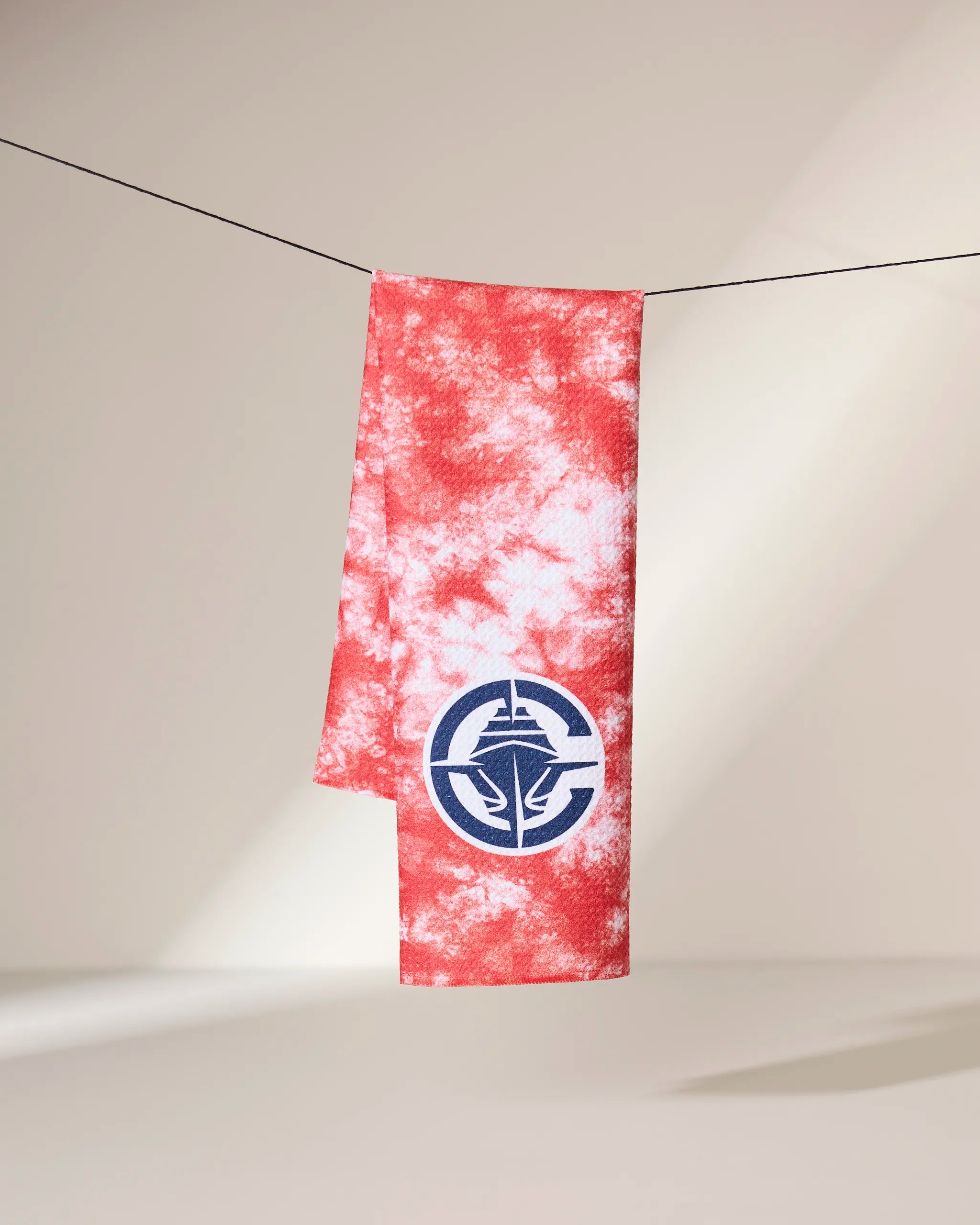 The Clipper Flash towel by Geometry, featuring a red and white tie-dye design with a blue nautical emblem, hangs on a black line against a neutral background.
