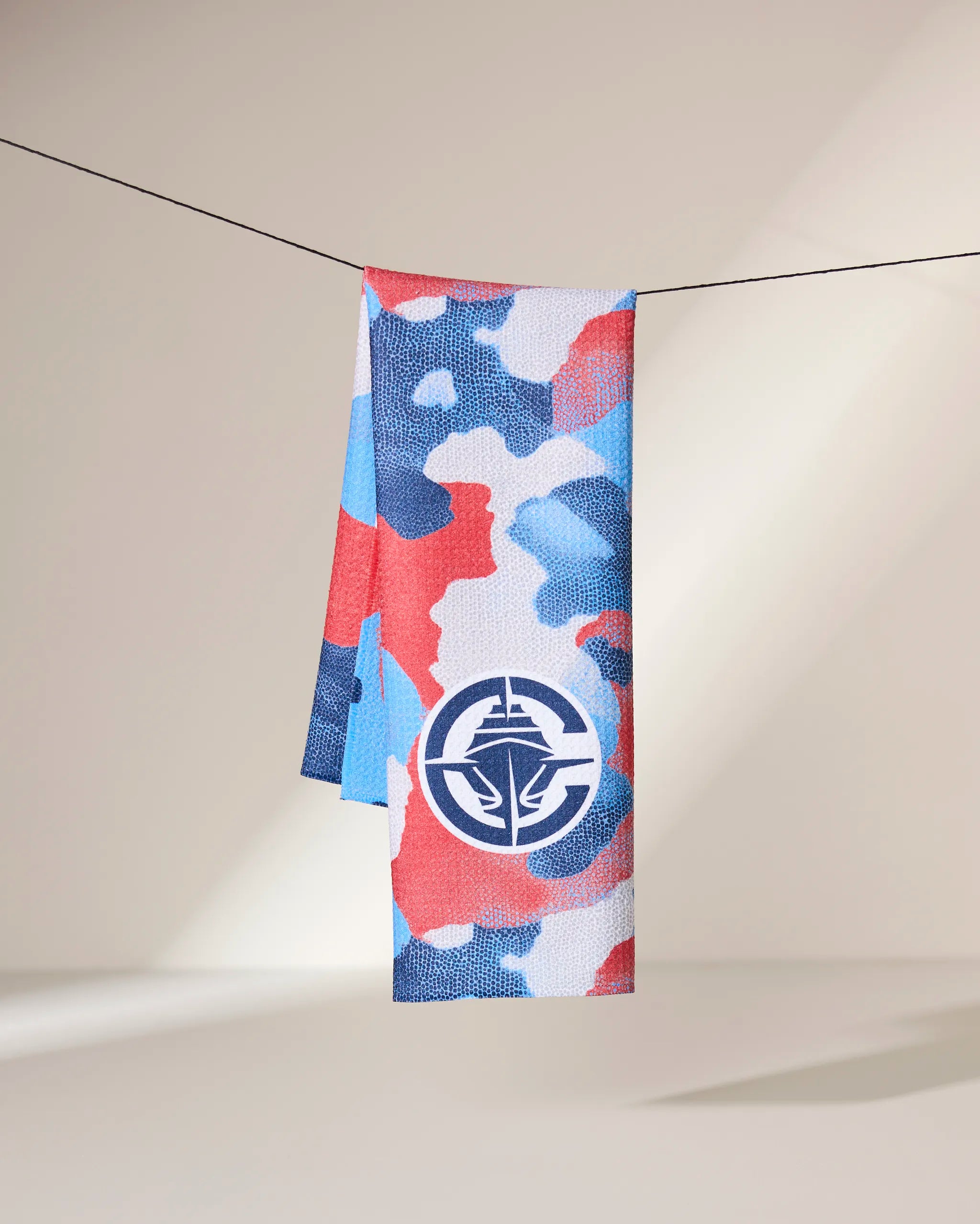 The Geometry Clippers Camo towel, featuring a red, blue, and white camouflage pattern and a bold circular logo near the bottom, hangs on a line against a neutral background with soft lighting.