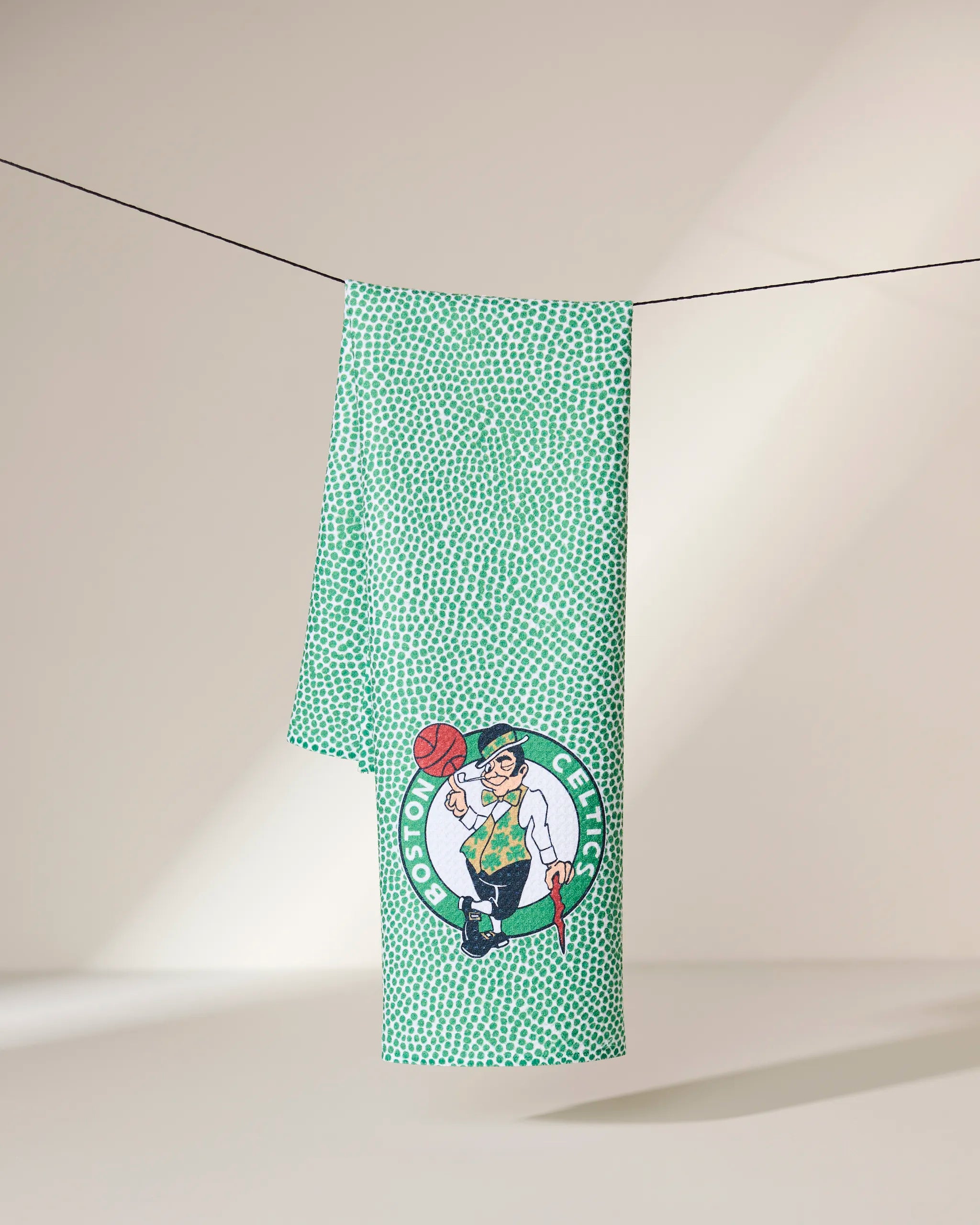 A green Celtics Grip towel by Geometry with a white dotted pattern hangs on a thin line, featuring the Boston Celtics logo of a leprechaun spinning a basketball against a light background.