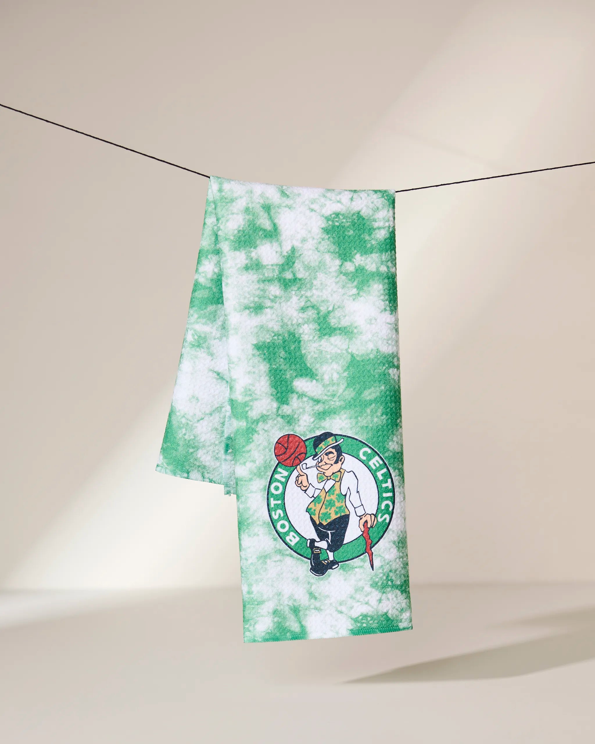 The Celtics Flash towel by Geometry, featuring a green and white tie-dye design with the Boston Celtics leprechaun logo, hangs on a clothesline against a neutral background.