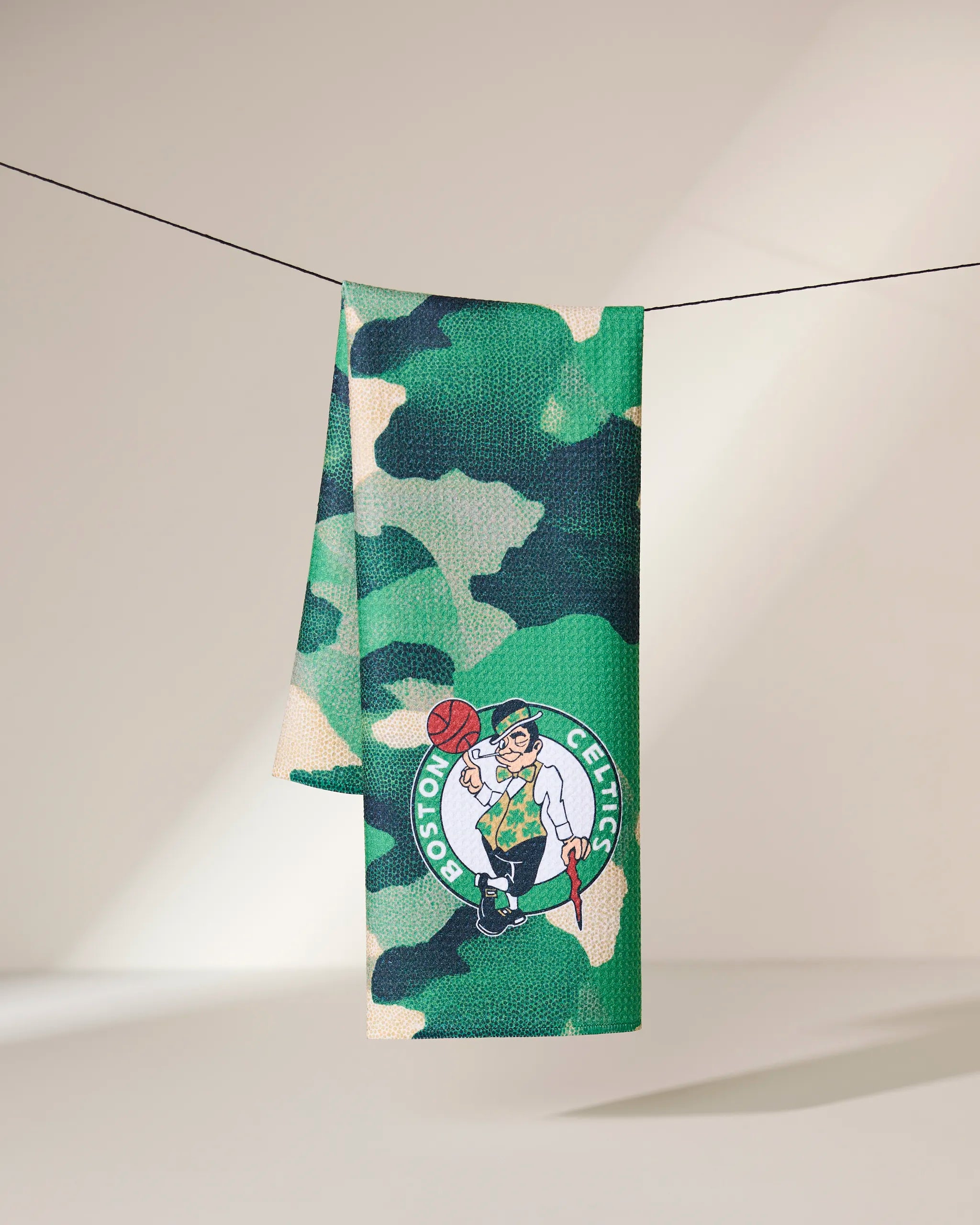 The Geometry Celtics Camo towel, featuring the Boston Celtics leprechaun logo on green camouflage, hangs on a black line against a softly shaded background.