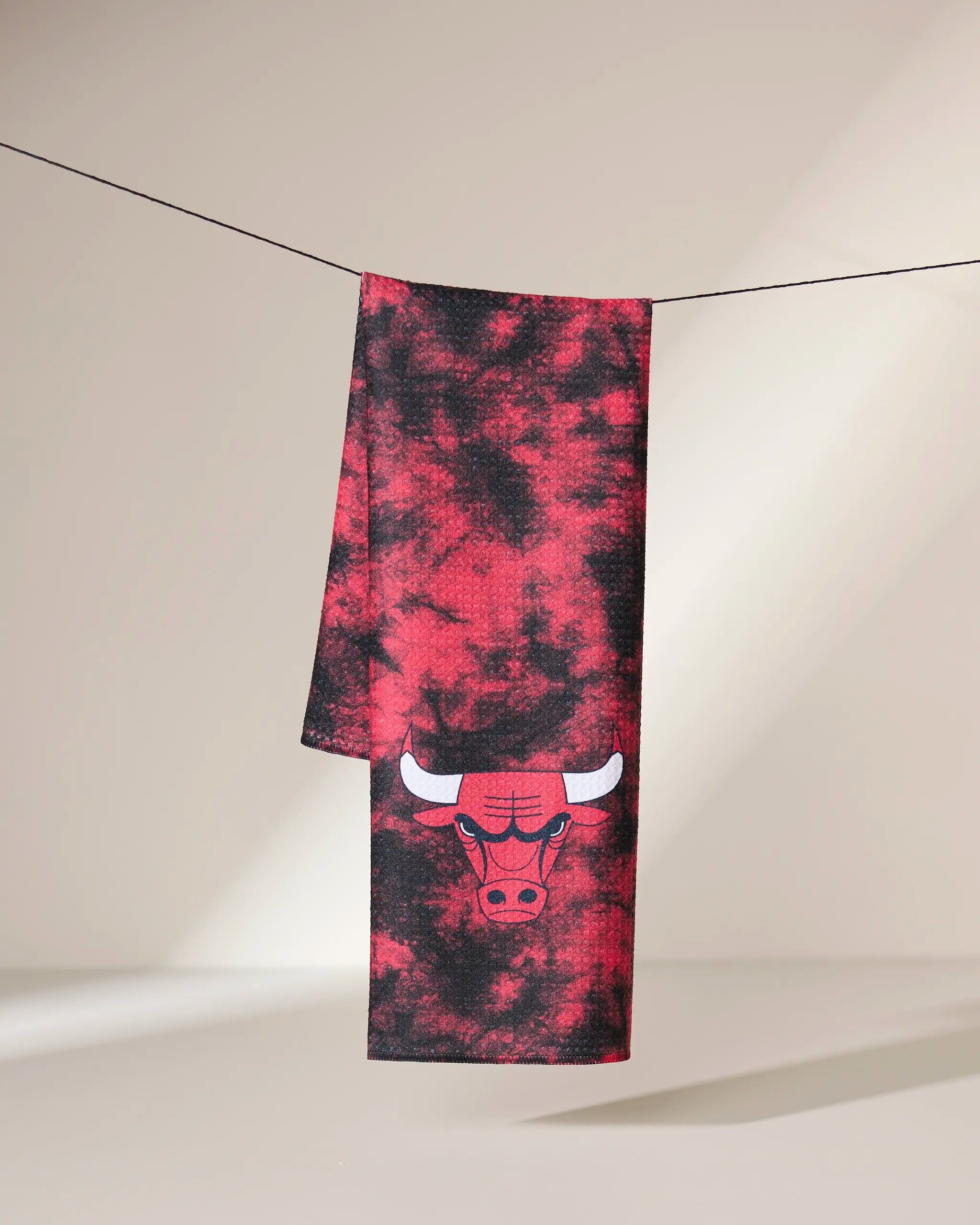 The Geometry Bulls Flash kitchen towel, in black and red tie-dye with the Chicago Bulls logo, hangs over a slim black wire against a neutral background.