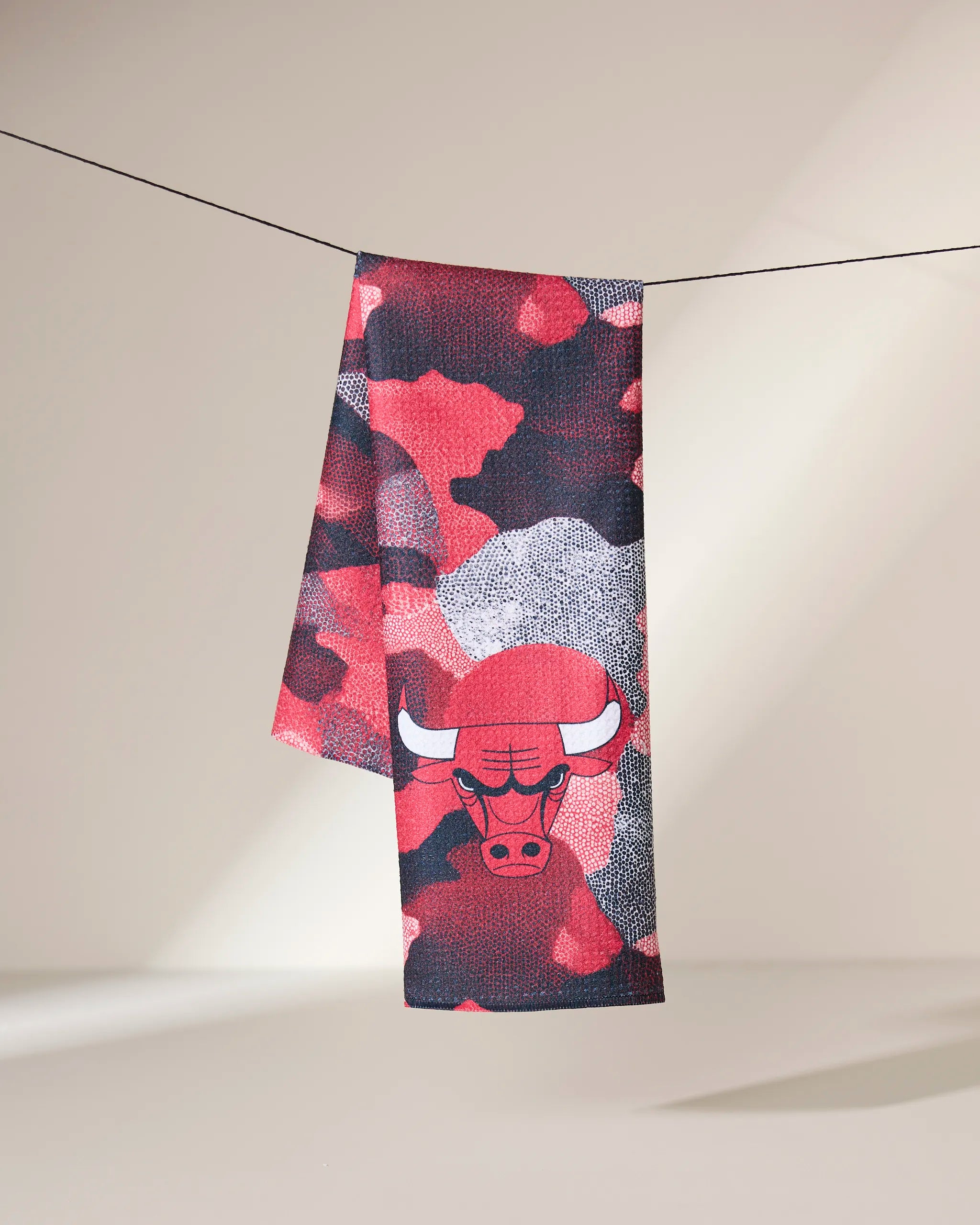 The Geometry Bulls Camo quick-drying towel, featuring a red, black, and gray camouflage pattern with the Chicago Bulls logo, hangs on a clothesline against a neutral background.