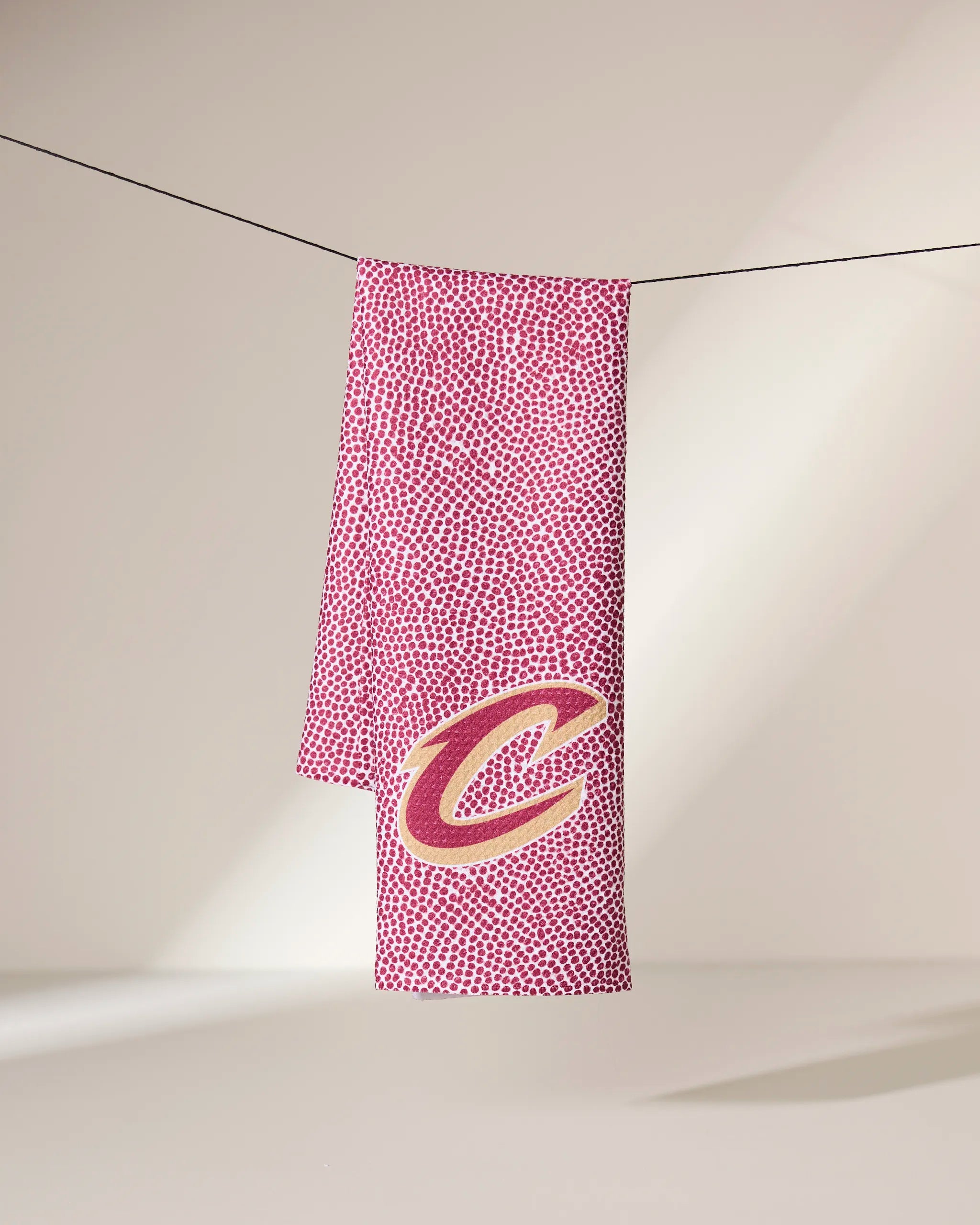 The Geometry Cavaliers Grip Red towel, featuring a red and white pattern with a large yellow and red letter C logo, hangs on a clothesline against a plain light background.