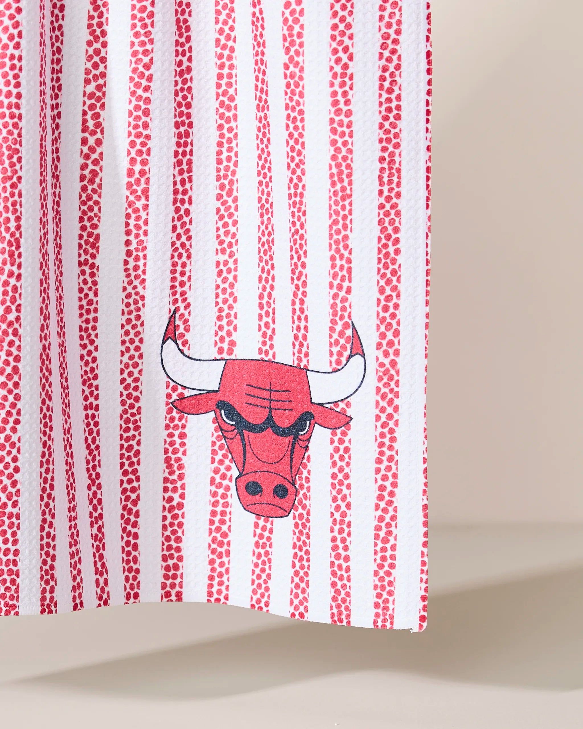 The Bulls Grip Stripe kitchen towel by Geometry is made from recycled materials and features white fabric with vertical red stripes and dotted lines, plus the Chicago Bulls logo near the bottom right corner.