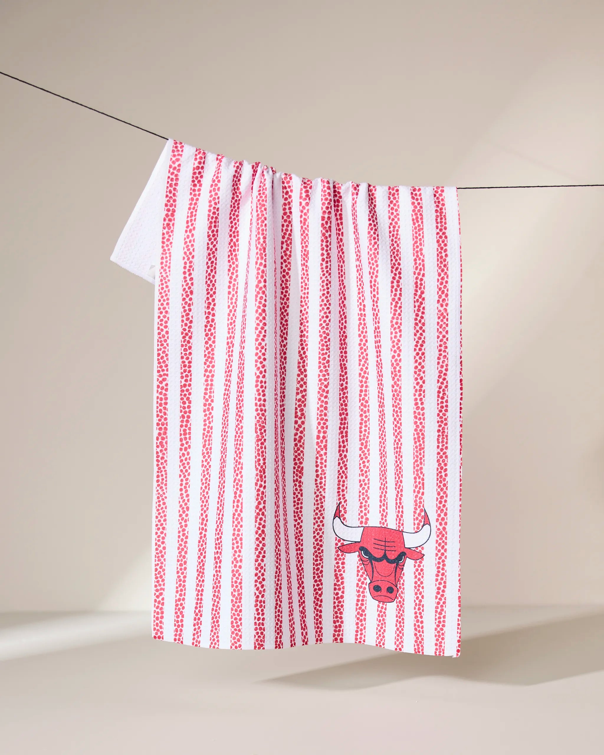 The Geometry Bulls Grip Stripe kitchen towel, featuring vertical red stripes and a red bull head logo, hangs on a clothesline against a beige background.
