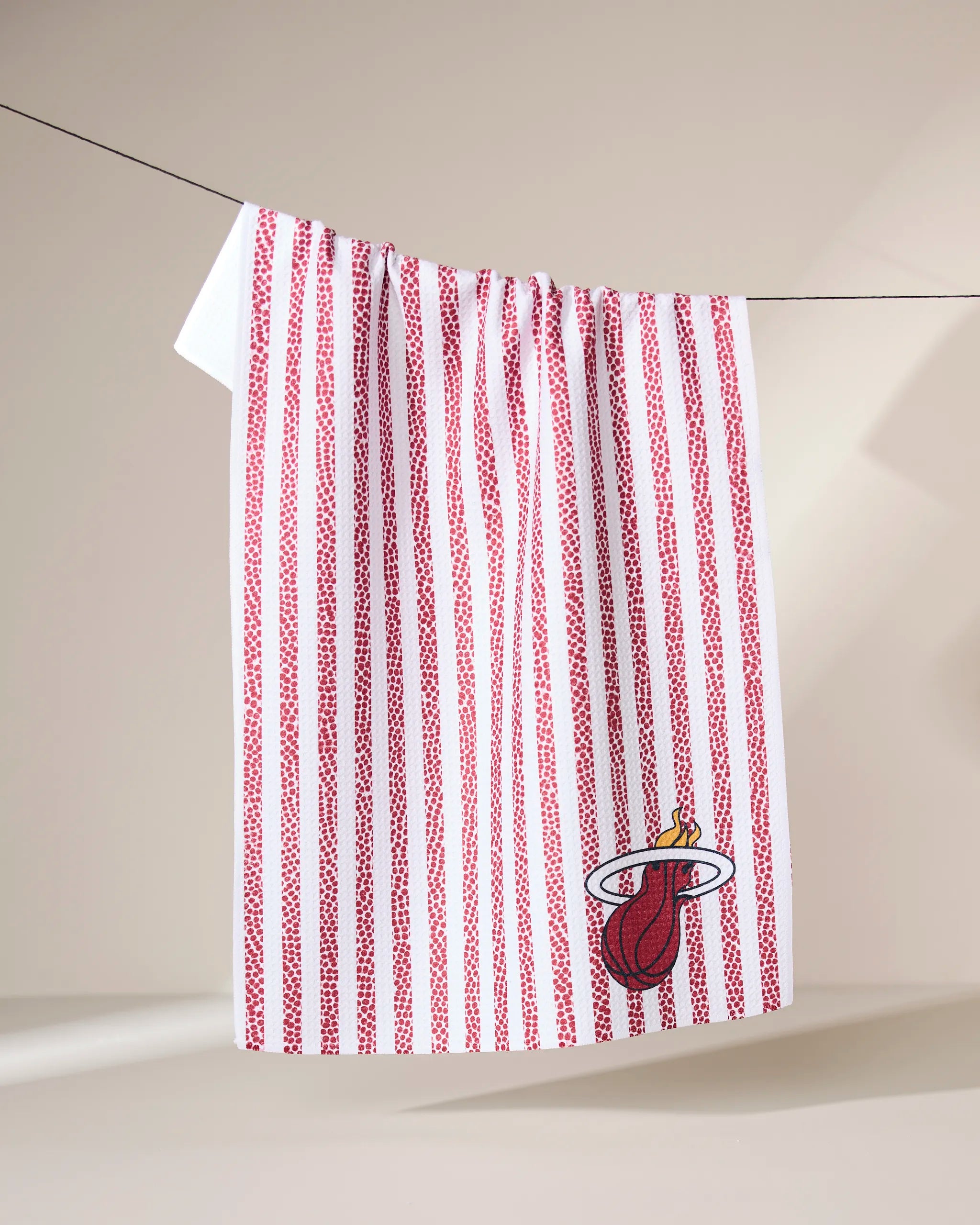 A Geometry Heat Grip Stripe Red towel with vertical red stripes and a Miami Heat logo hangs on a clothesline against a softly lit neutral background.