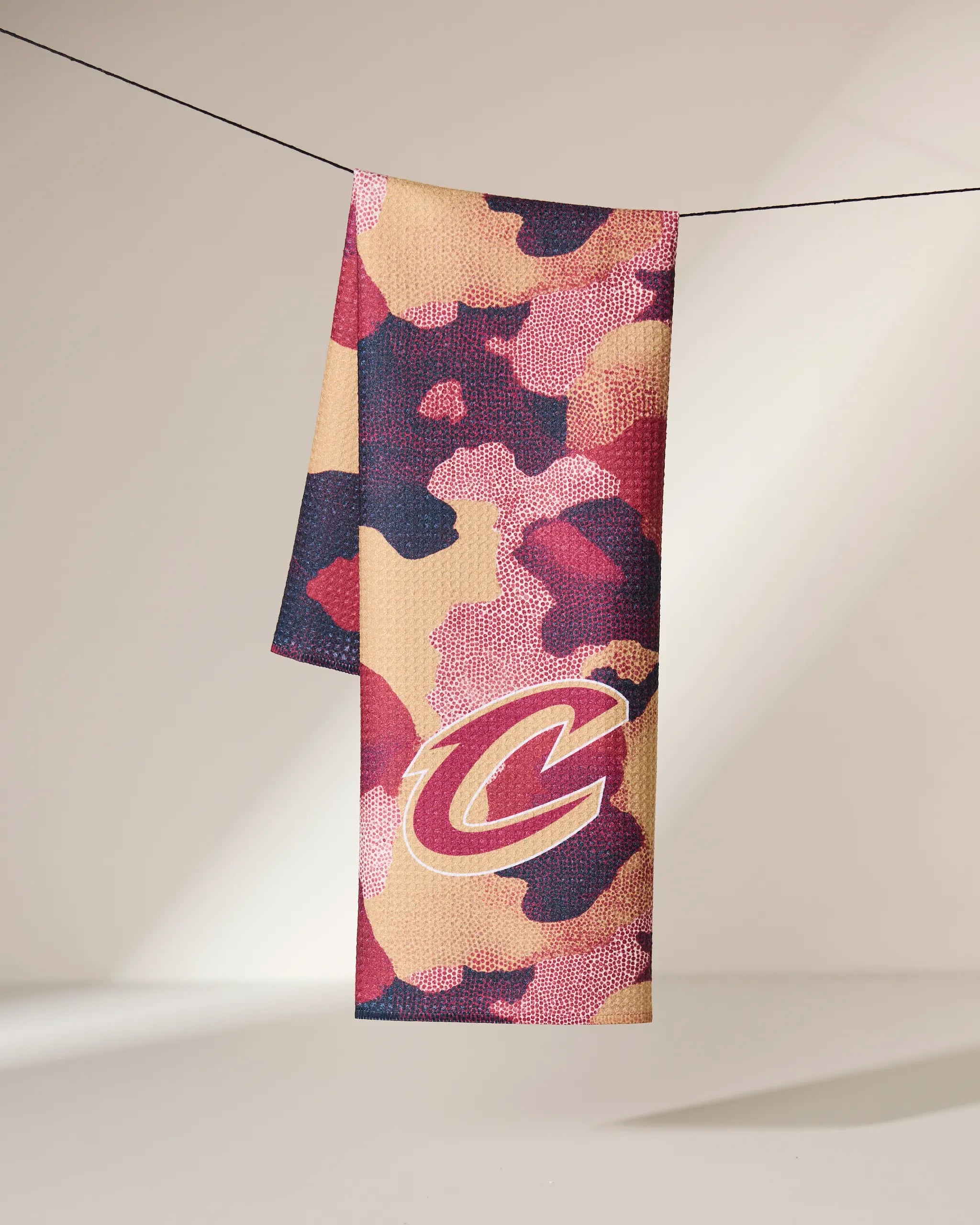 The Geometry Cavaliers Camo recycled kitchen towel features a red, maroon, pink, and tan camouflage pattern with a large C logo, hanging over a black line against a neutral background.