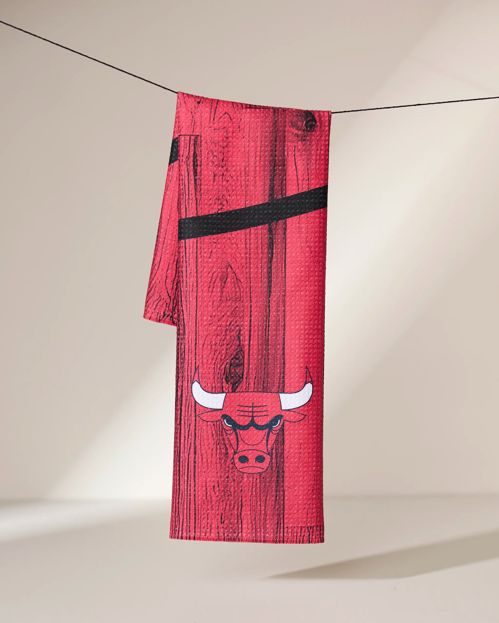 The Geometry Bulls Zone towel, featuring a red ultra-absorbent fabric with a wood-grain pattern and white bulls head logo, is displayed hanging over a black string against a neutral background.
