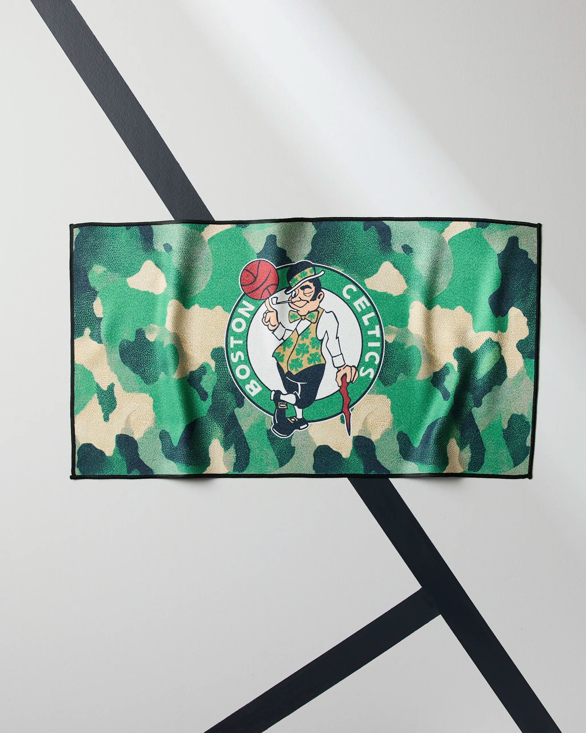 The Celtics Camo by Geometry is a rectangular NBA fitness towel with a green camouflage pattern and the Boston Celtics leprechaun logo, shown on a white backdrop with black diagonal lines.