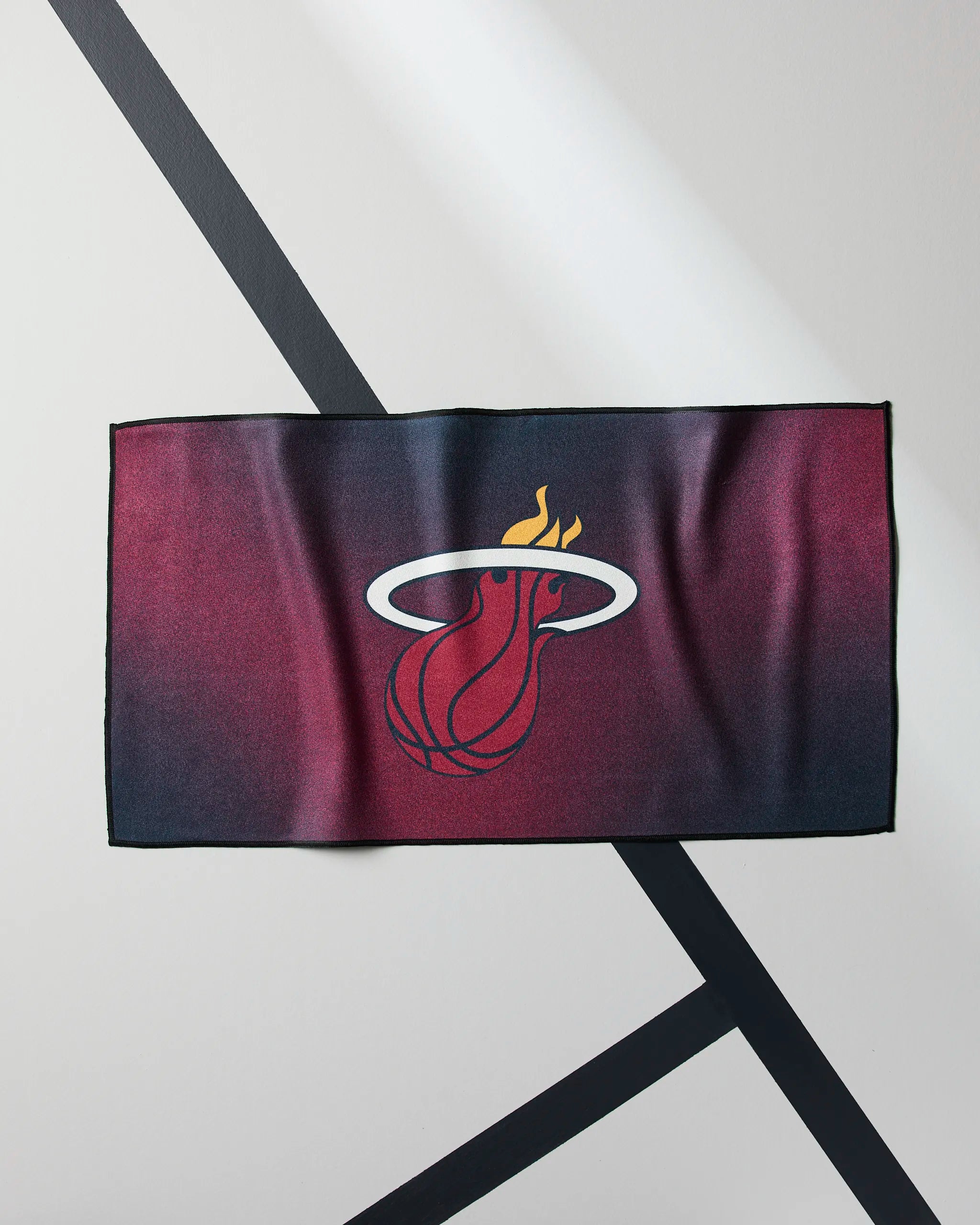 The Geometry Heat Fade towel is a rectangular NBA fitness towel with the Miami Heat logo—a flaming basketball through a white hoop—on a red and black gradient, displayed on a diagonally striped light surface.