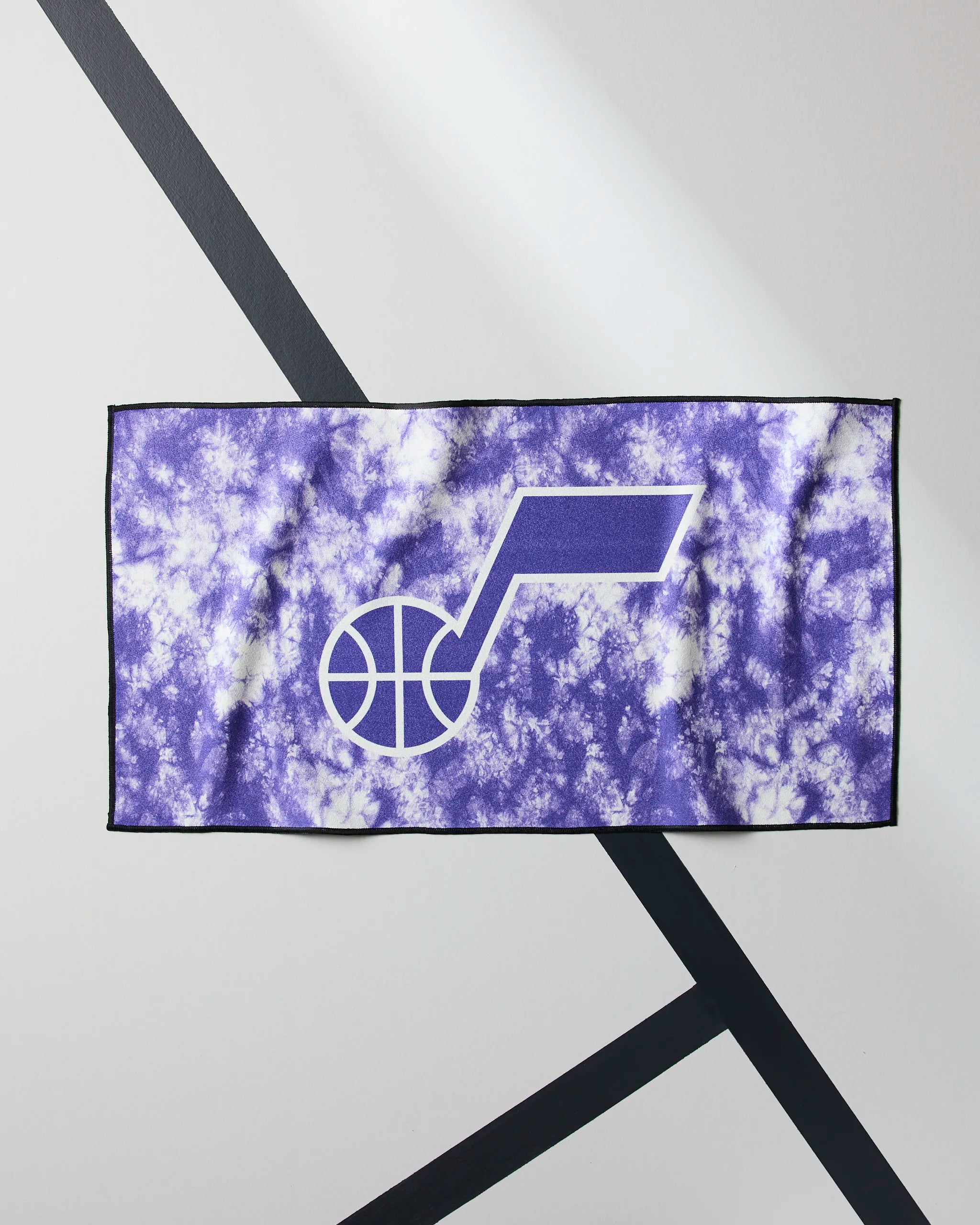 The Jazz Flash by Geometry is a purple and white tie-dye NBA fitness towel with a bold white Utah Jazz note logo, displayed flat on a gray surface with black crisscross diagonal lines.