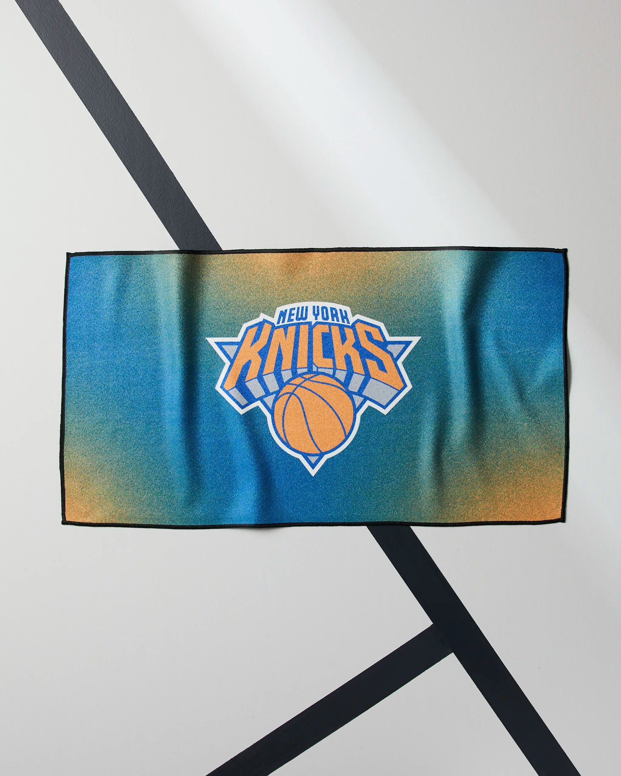 The Knicks Fade by Geometry is a rectangular blue and orange NBA fitness towel featuring the New York Knicks logo with a basketball and text, shown on a white surface with black diagonal lines.