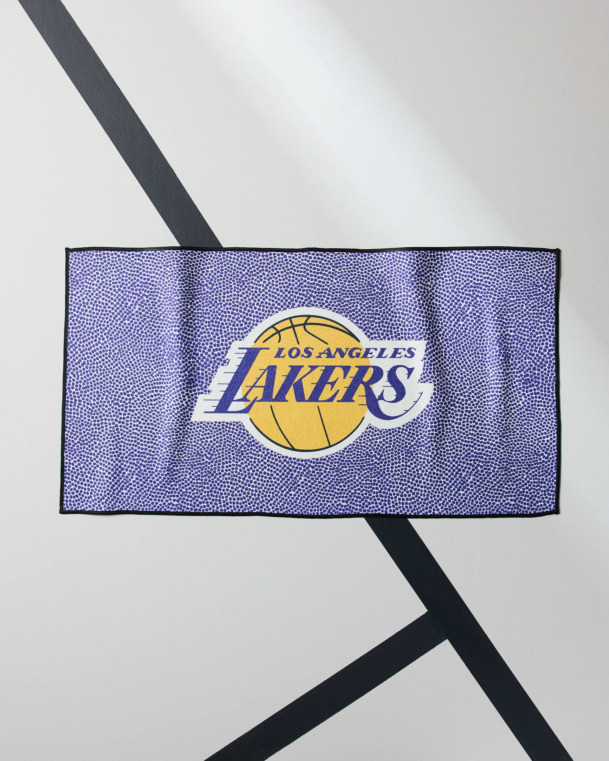 The Geometry Lakers Grip is a rectangular NBA fitness towel featuring the Los Angeles Lakers logo with a yellow basketball and purple text, displayed on a white floor with intersecting black lines.