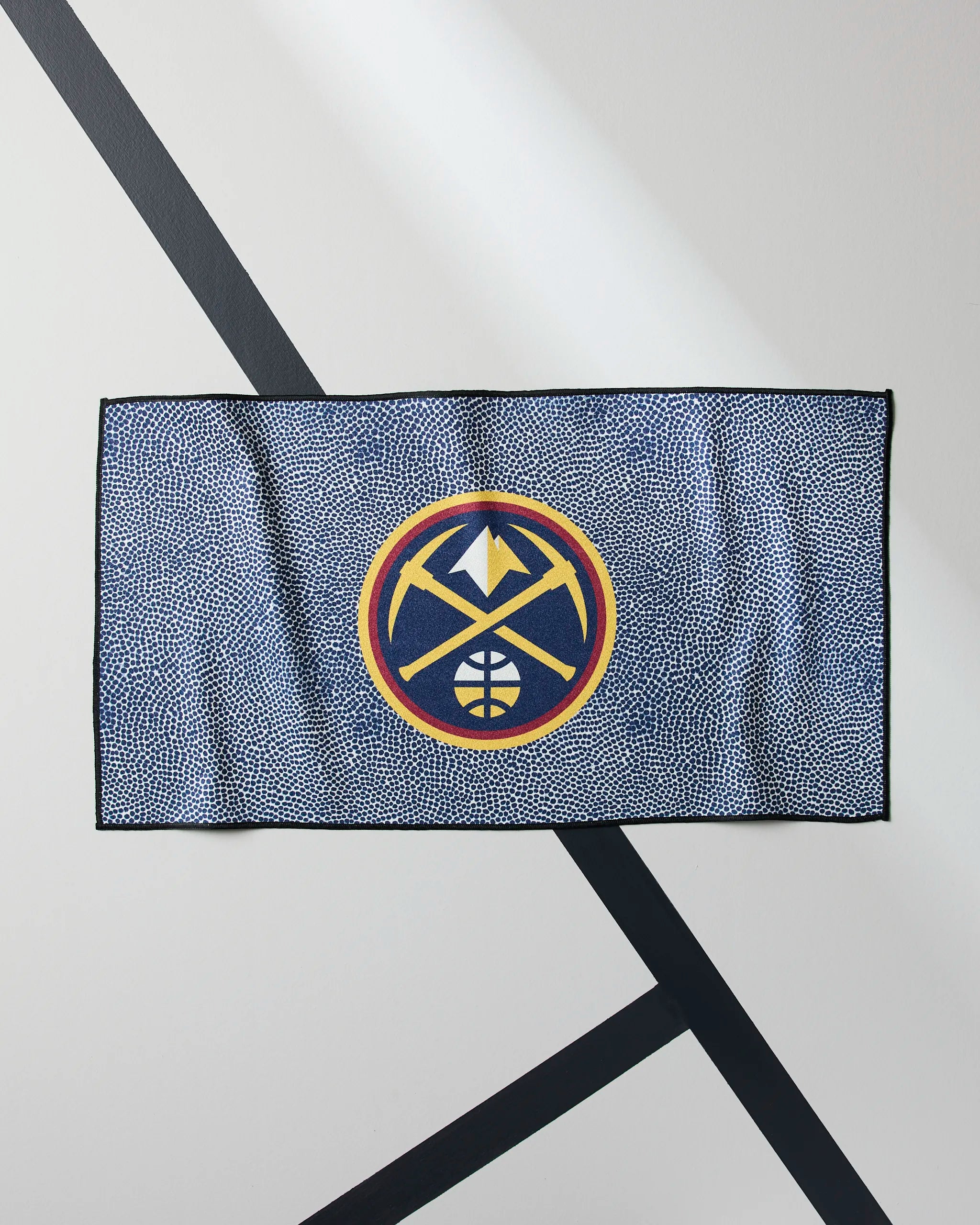 The Nuggets Grip by Geometry is a blue NBA fitness towel with the Denver Nuggets logo in yellow, navy, and white. Displayed on a white wall with black diagonal lines, it's perfect as a quick-drying gym towel for fans and athletes.