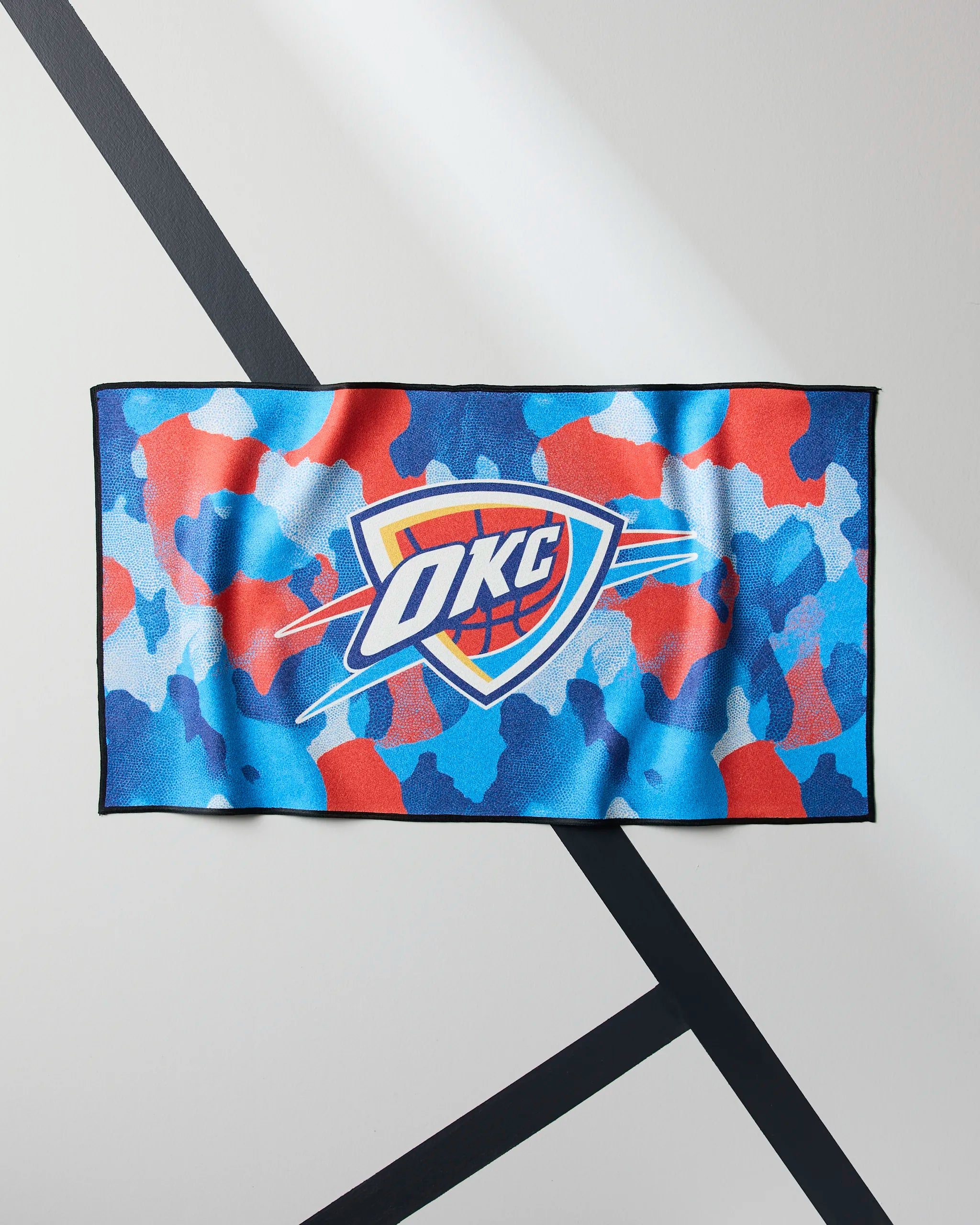 The OKC Camo by Geometry is a rectangular quick-drying gym towel with a blue, red, and white camouflage pattern and the OKC Thunder logo in the center, shown hanging against a wall with black diagonal lines.