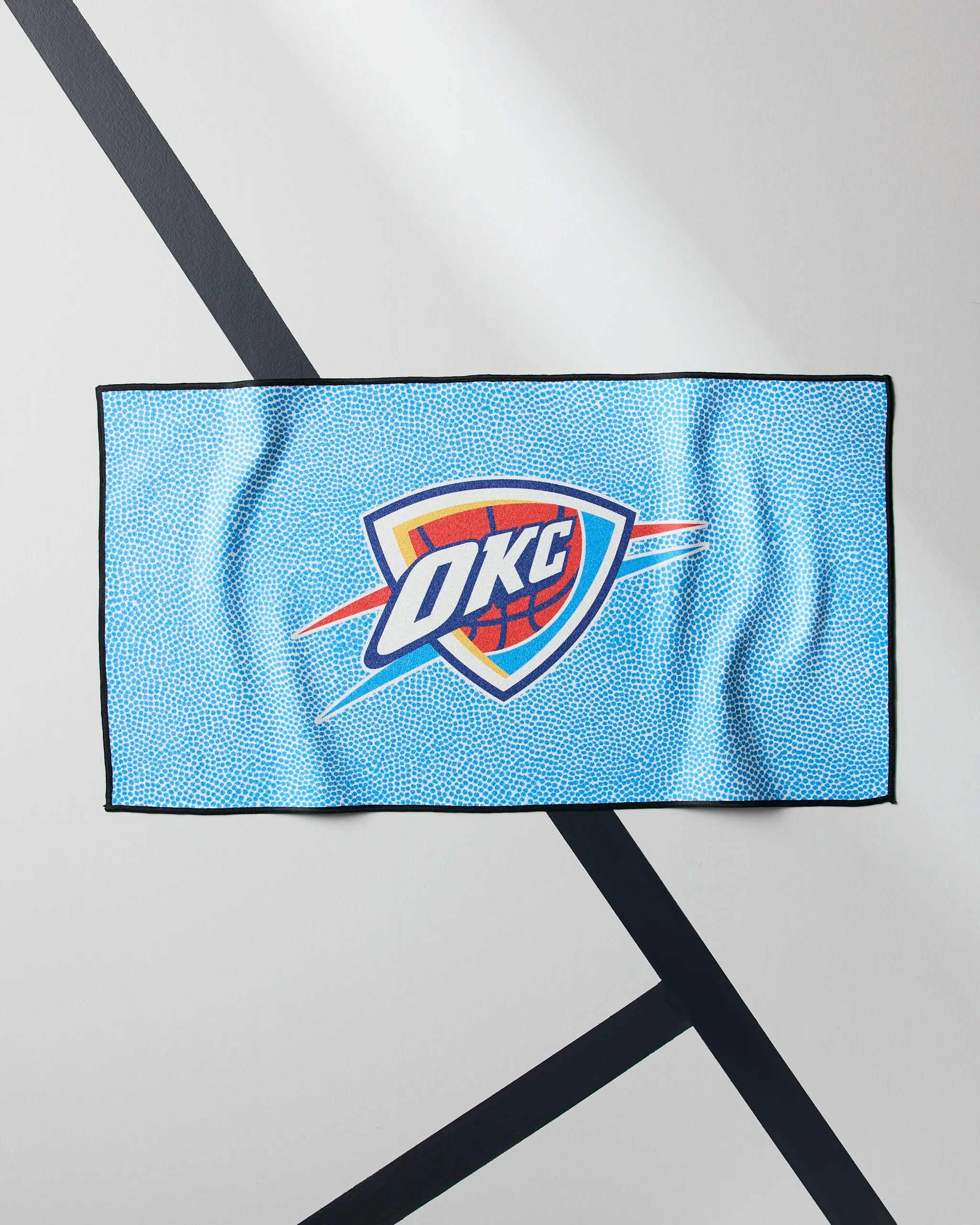 A light blue Geometry OKC Grip fitness towel with the Oklahoma City Thunder logo is shown lying flat on a white surface featuring black diagonal lines in the background.