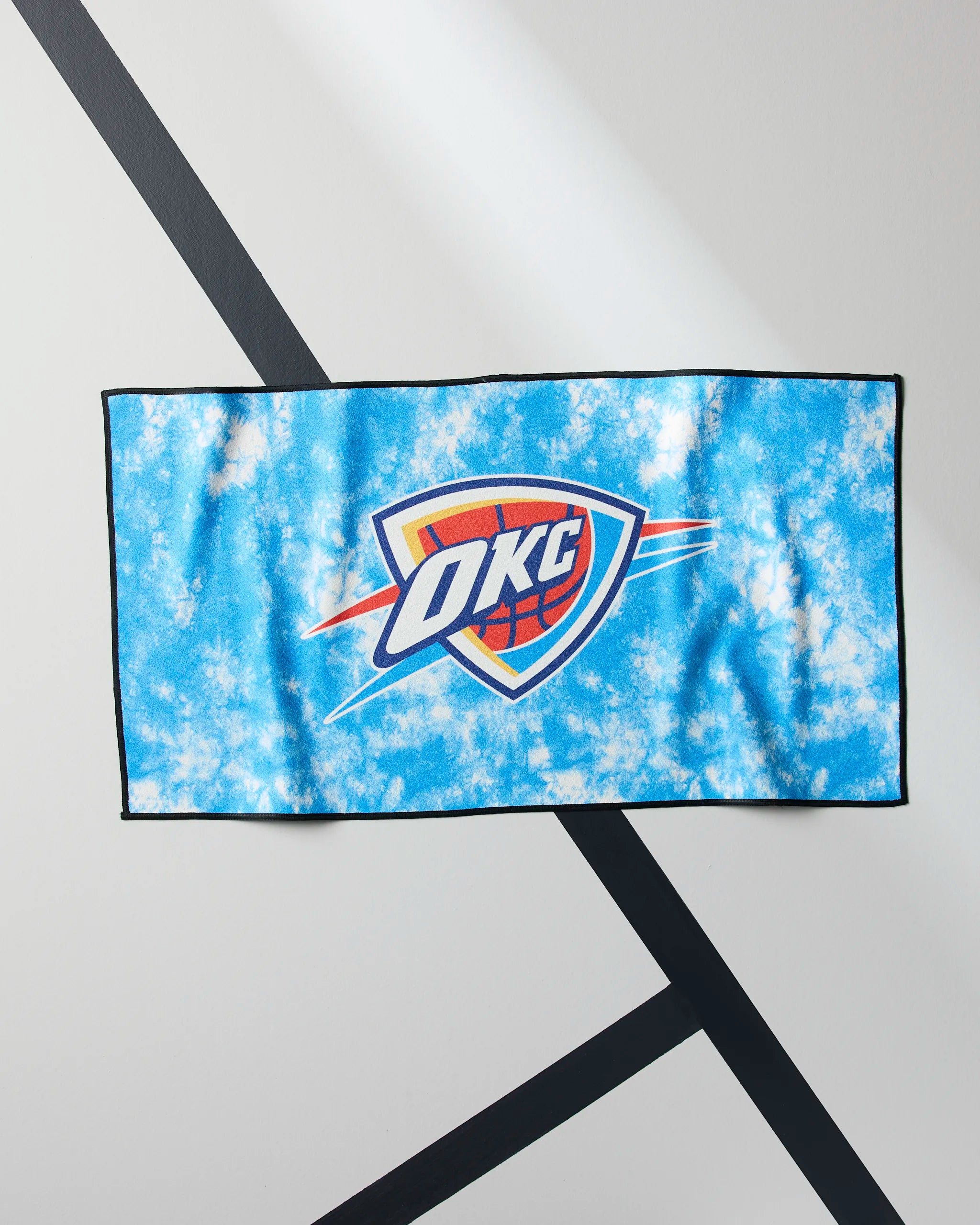 The Geometry OKC Flash is a blue and white tie-dye NBA fitness towel featuring the Oklahoma City Thunder logo, displayed on a white wall with black geometric lines.