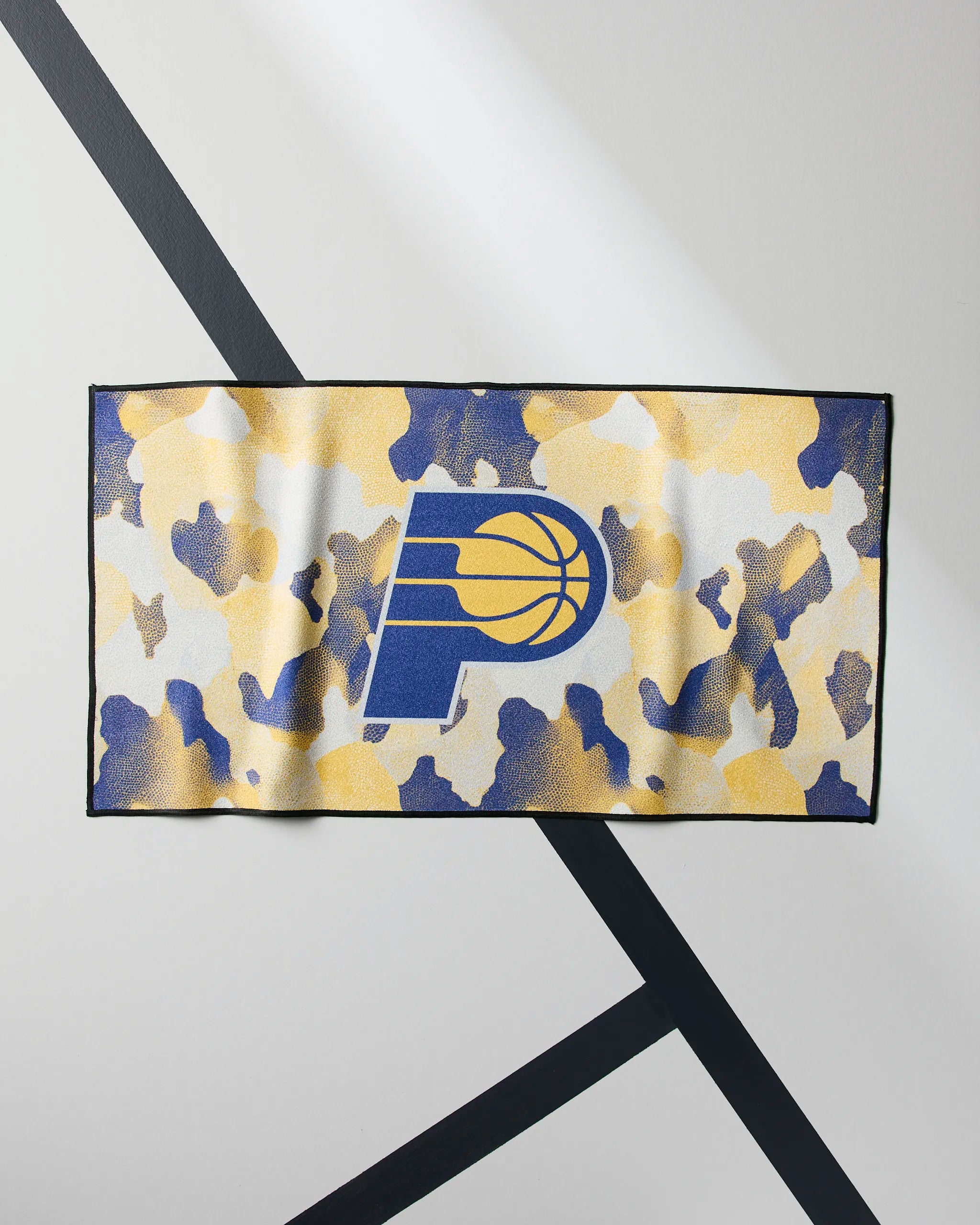 The Geometry Pacers Camo towel is a rectangular NBA fitness towel with a blue and yellow camouflage pattern and the Indiana Pacers logo centered, shown on a white surface with black diagonal lines.