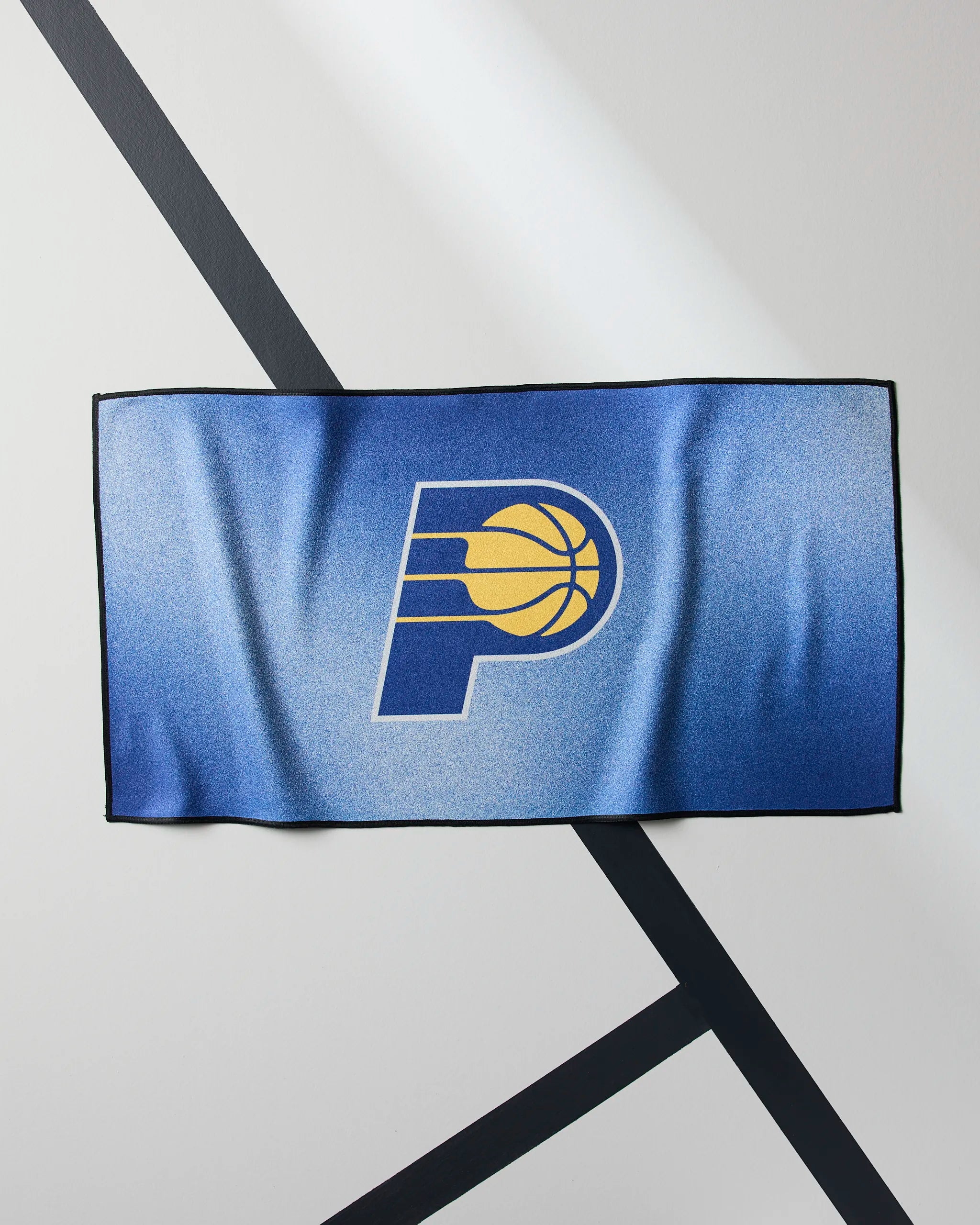 The Geometry Pacers Fade is a blue NBA fitness towel with the Indiana Pacers logo, shown on a white, diagonally-striped surface—ideal for quick-drying use at the gym or during intense workouts.