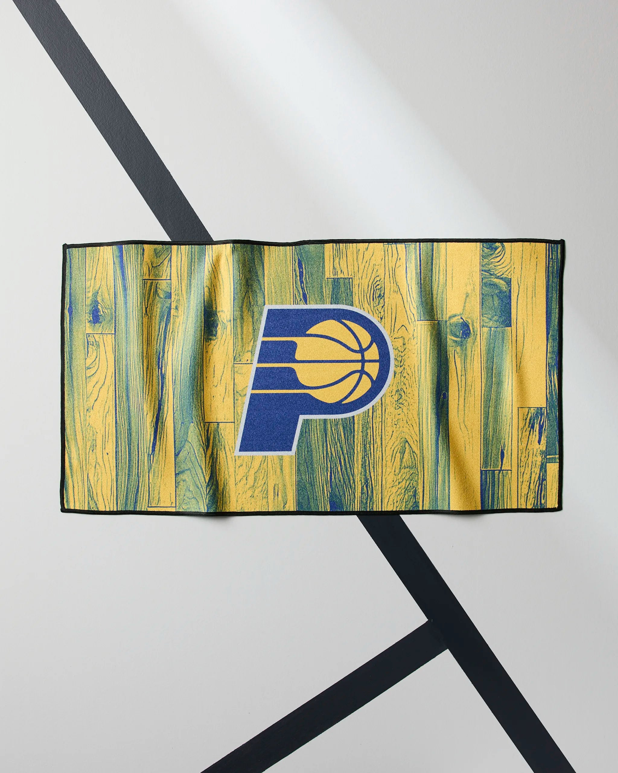 The Pacers Zone towel by Geometry is rectangular with a yellow and green woodgrain design and features the Indiana Pacers logo in blue and gold at the center. It’s shown flat on a geometric, light-colored surface.