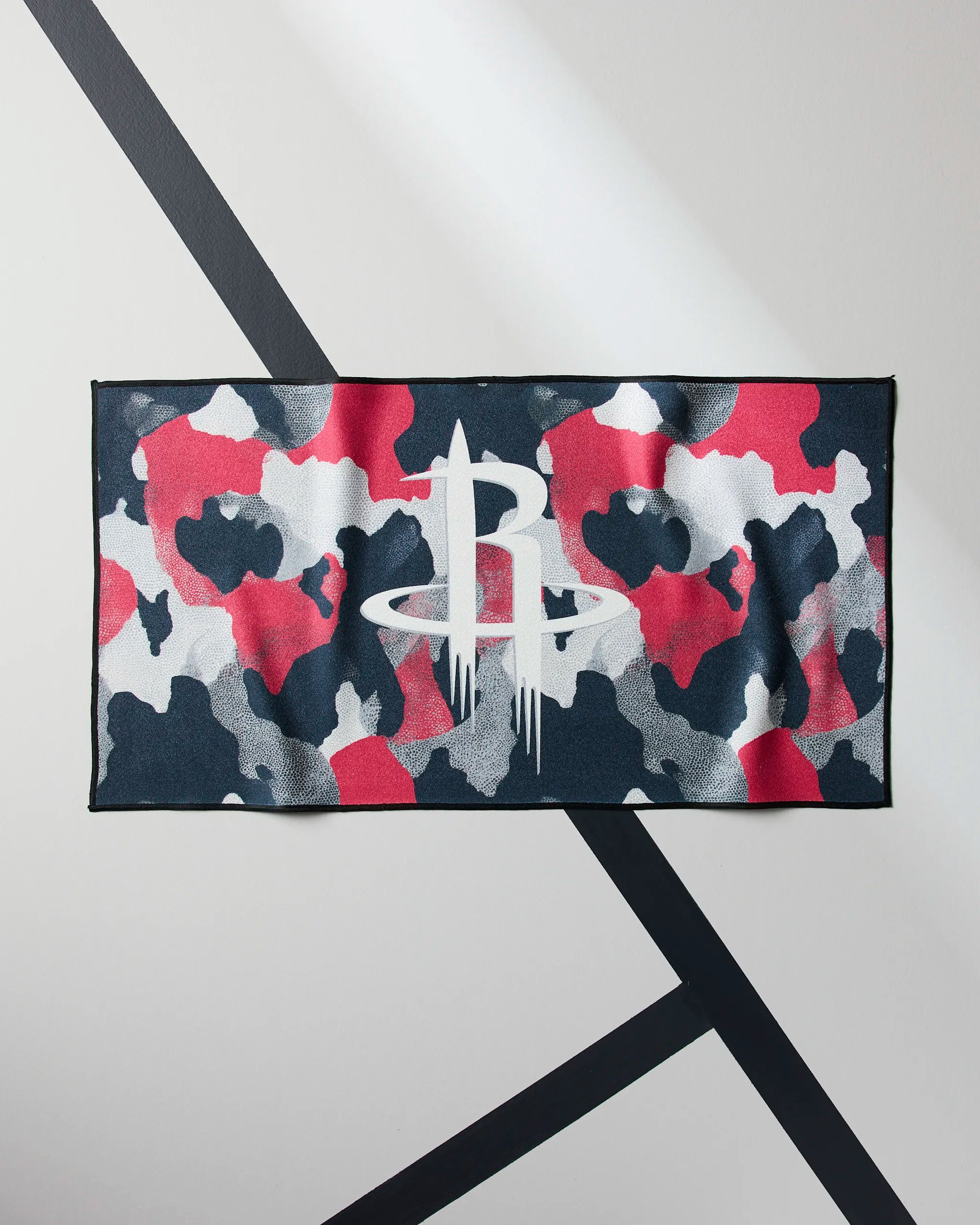 The Rockets Camo fitness towel by Geometry, featuring a red, gray, black, and white camouflage design with a bold white Houston Rockets logo at the center, is shown displayed on a diagonally striped wall.