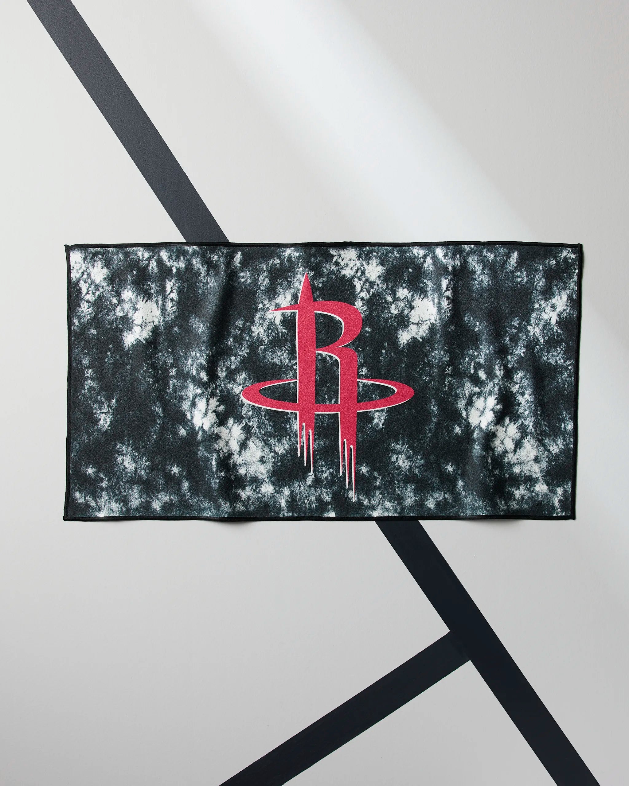 The Geometry Rockets Flash towel features a black and white tie-dye design with a bold red Houston Rockets logo at the center, showcased on a gray surface with black diagonal lines.