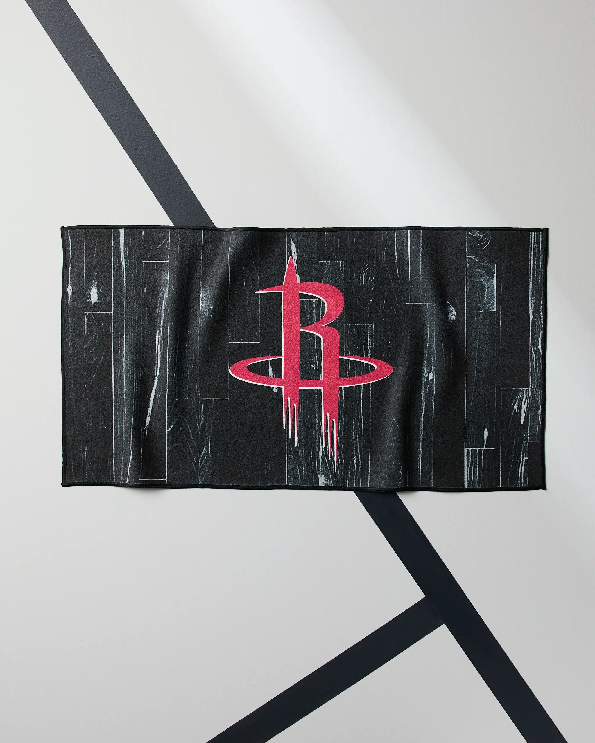 The Geometry Rockets Zone fitness towel, featuring a red Houston Rockets logo at the center on a black background, is shown laid flat on a light gray surface with intersecting black lines.
