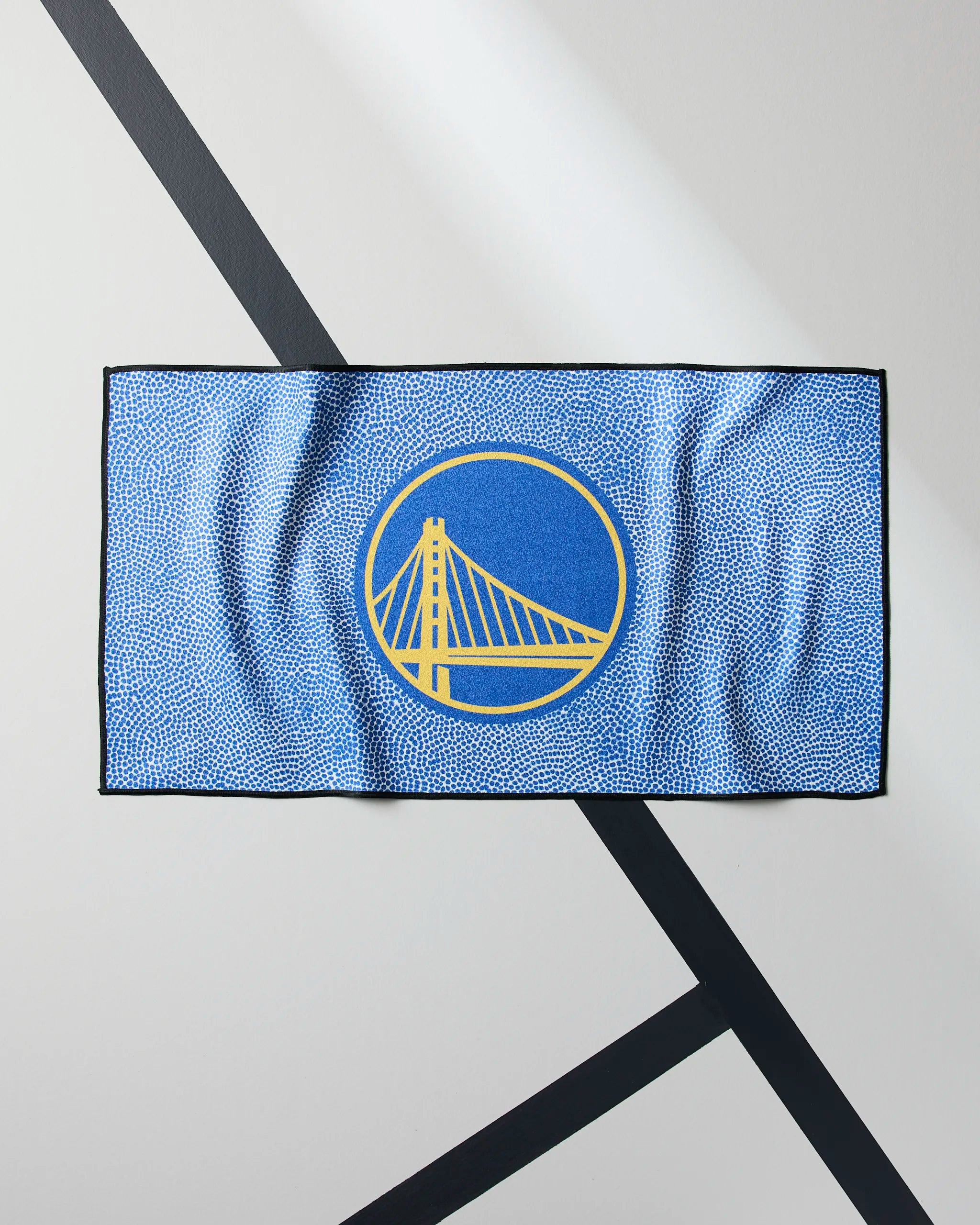 The Geometry Warriors Grip is a blue quick-dry workout towel with a yellow Golden State Warriors logo—featuring a stylized bridge in a circle—shown flat on a white surface with diagonal black lines.