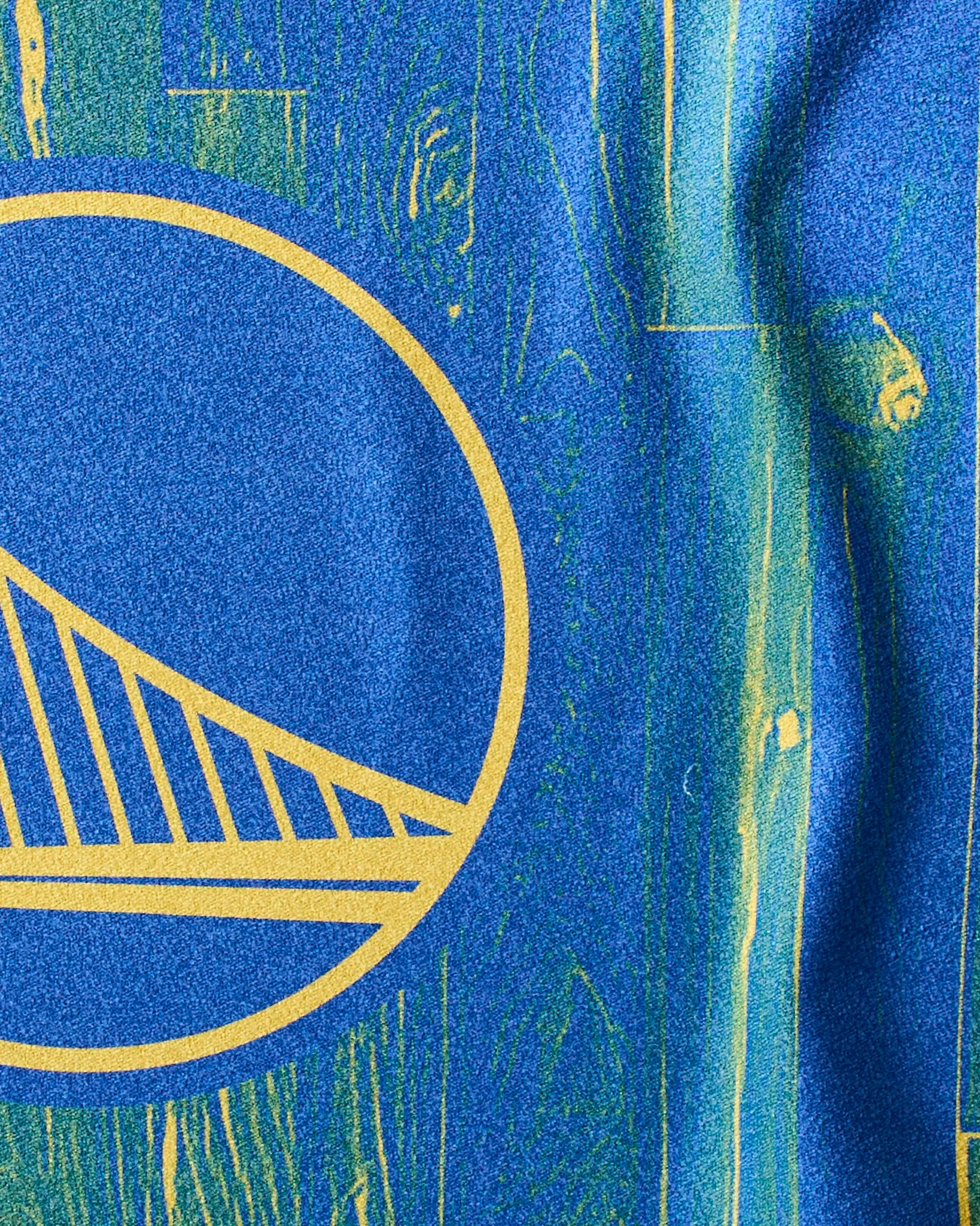 Close-up of the Warriors Zone NBA fitness towel by Geometry, showing part of the circular Golden State Warriors logo in yellow on a blue and yellow wood-grain textured background. The towel appears on the left side of the image.