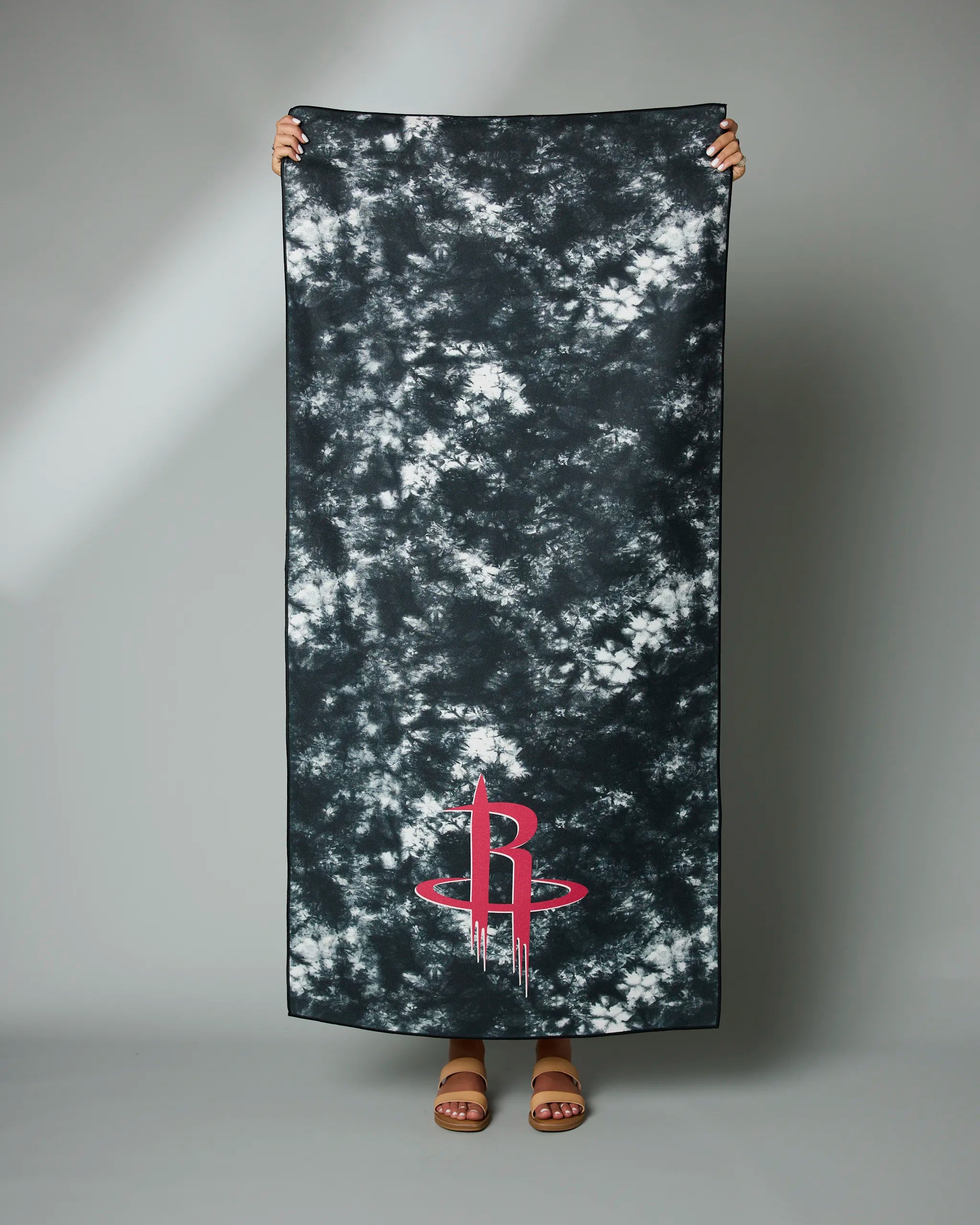 A person holds the Geometry Rockets Flash black and white tie-dye towel with a red Houston Rockets logo near the bottom, standing against a plain gray background.