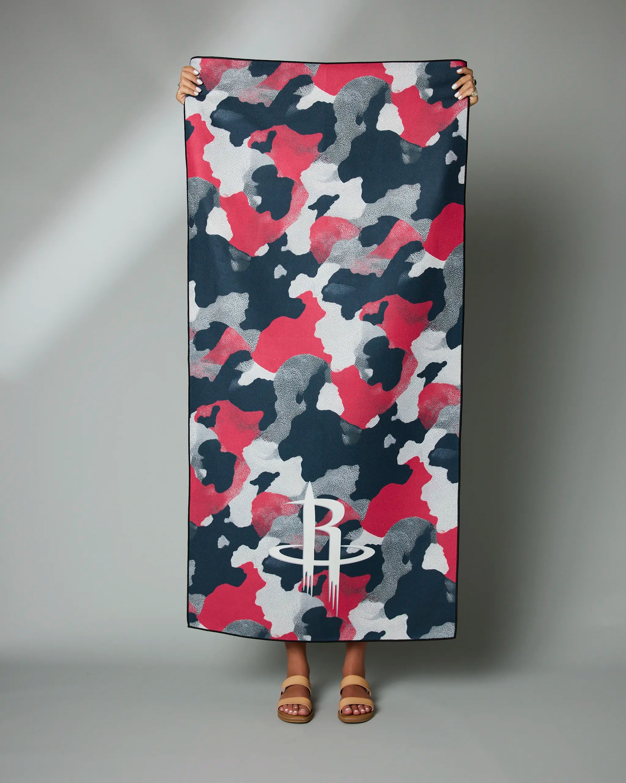 A person holds the Geometry Rockets Camo towel, featuring a rectangular shape with red, black, white, and gray camouflage and a white logo at the bottom. Only their hands and sandal-clad feet are visible.