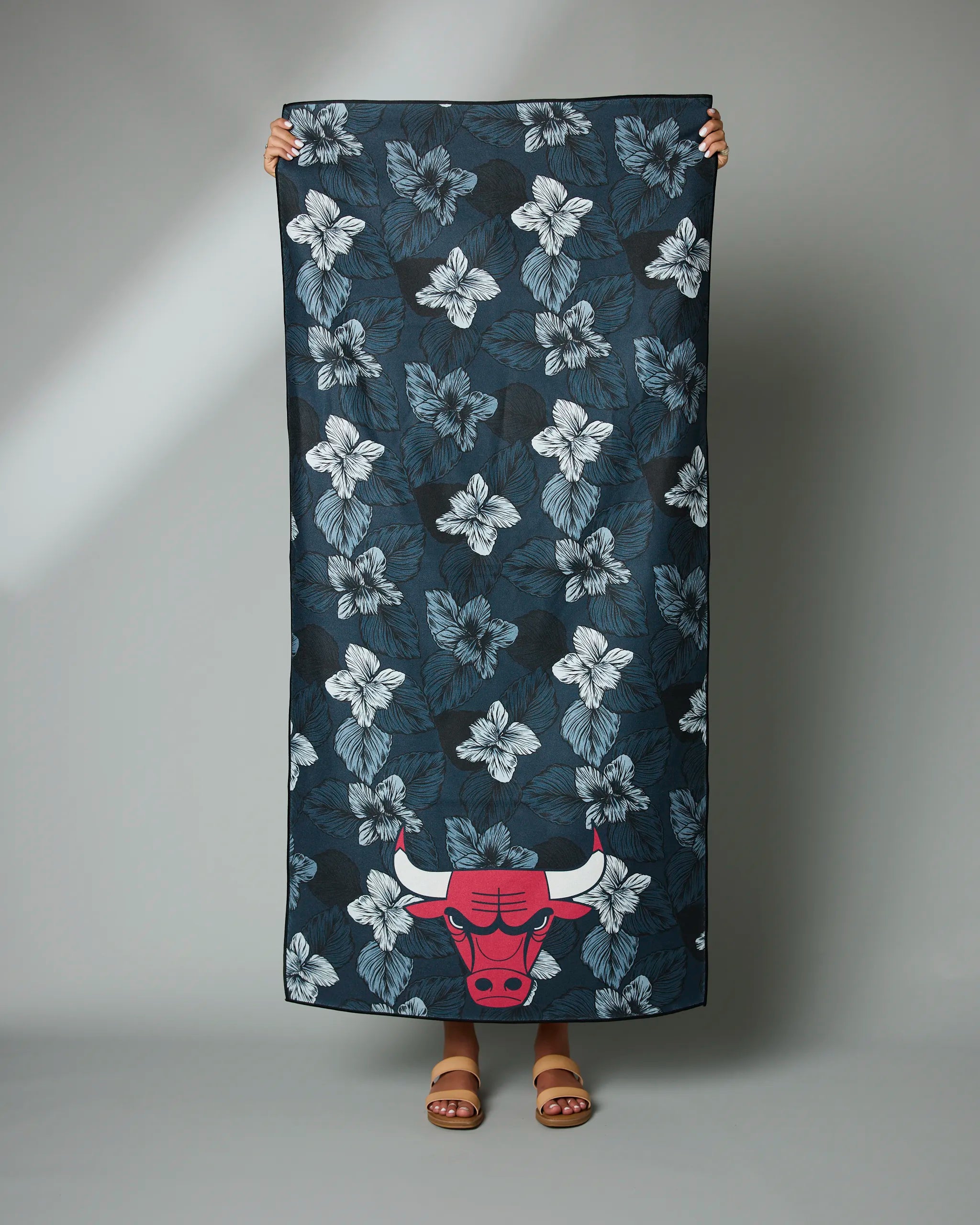 A person holds the Geometry Bulls Floral towel, featuring a dark floral design with white hibiscus flowers and a red Chicago Bulls logo at the bottom; only their hands and feet in sandals are visible.