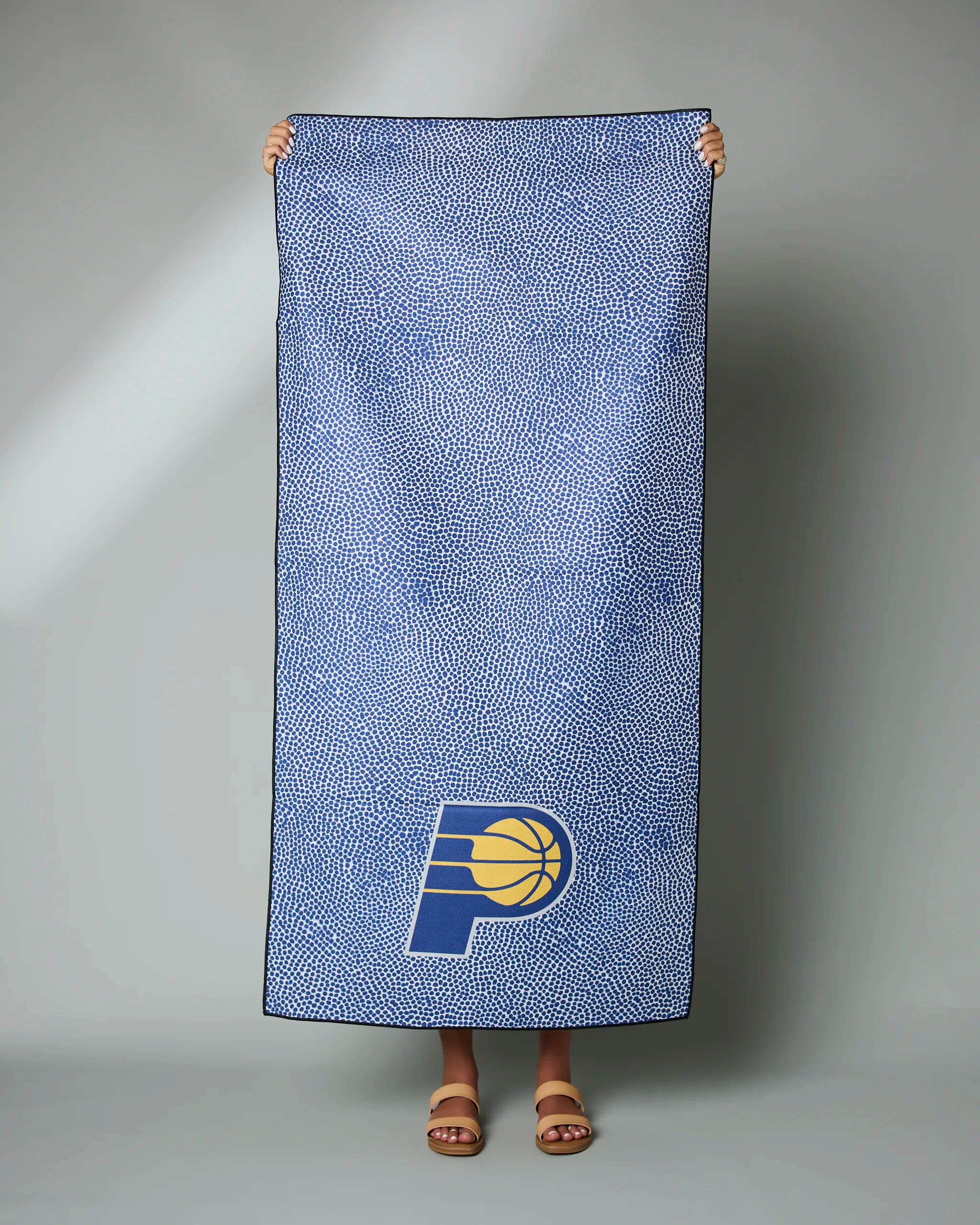A person is holding up the Geometry Pacers Grip towel, featuring a blue design with white speckles and the Indiana Pacers team logo at the bottom; only their hands and sandal-clad feet are visible behind it.