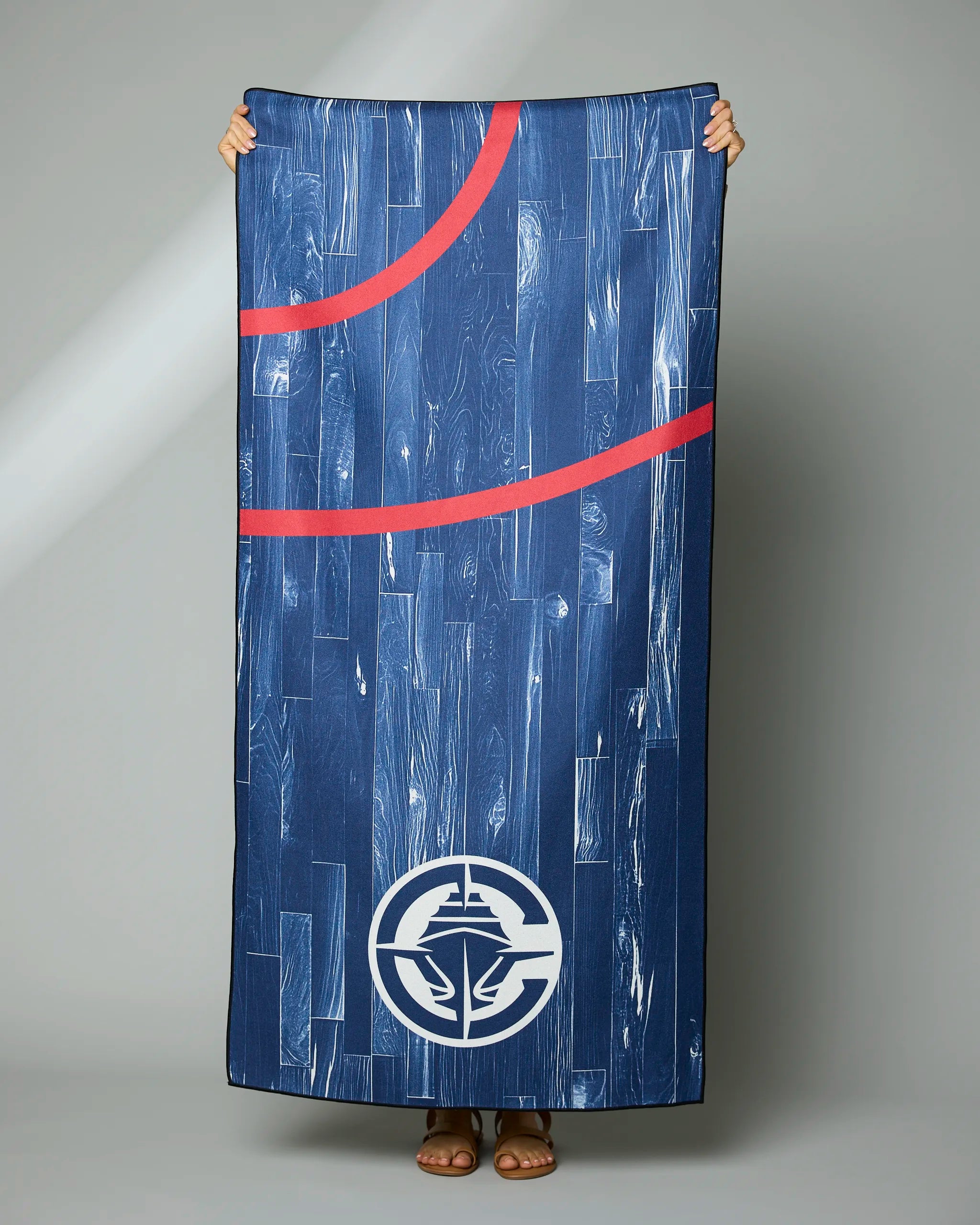 A person holds the Geometry Clippers Zone towel, featuring a wood grain pattern, two curved red lines, and the LA Clippers logo at the bottom. Only their hands, feet, and sandals are visible.