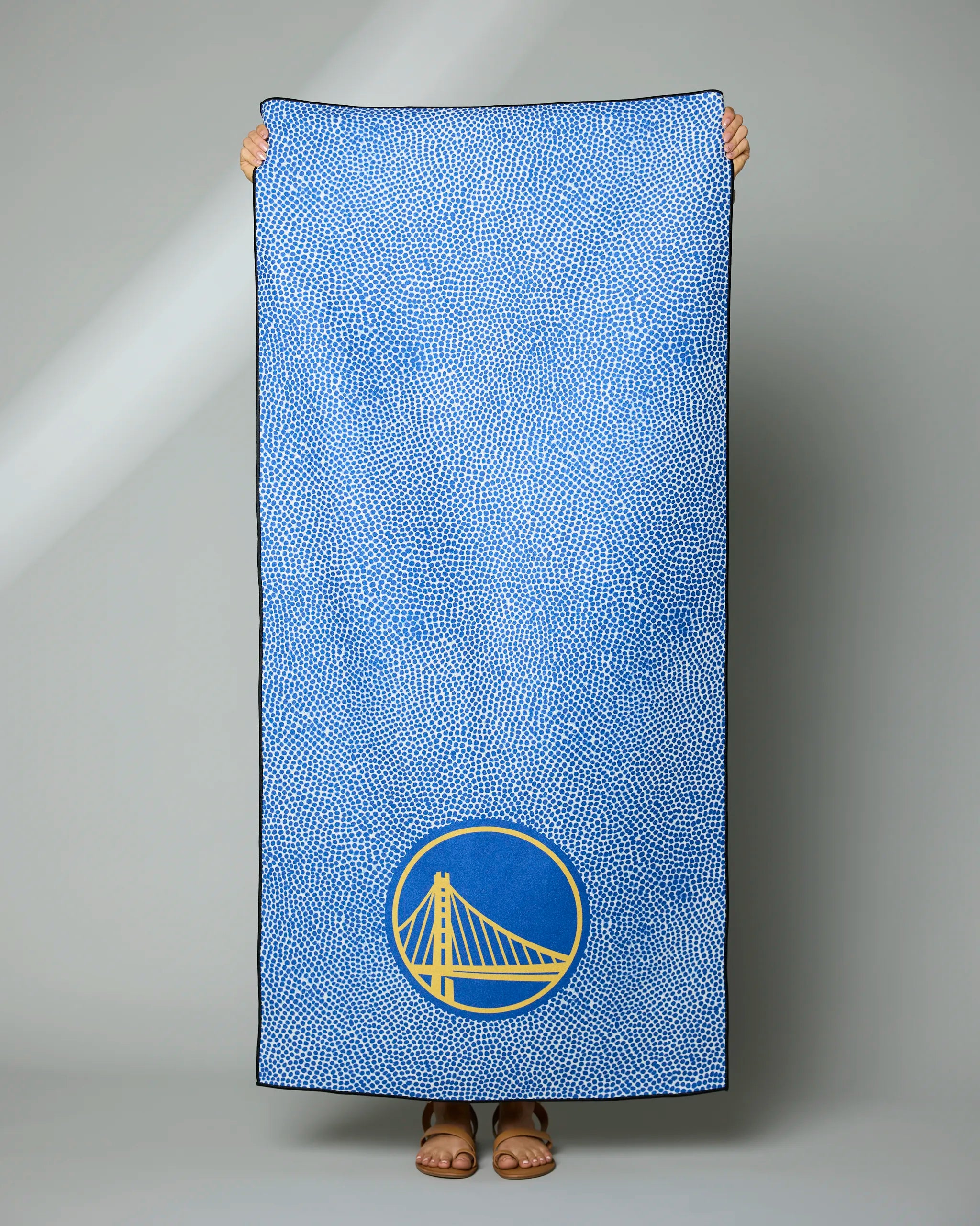 A person holds up the Geometry Warriors Grip towel, featuring a blue design with small white dots and a yellow Golden Gate Bridge logo in a blue circle near the bottom. Only their feet and hands are visible behind the towel.