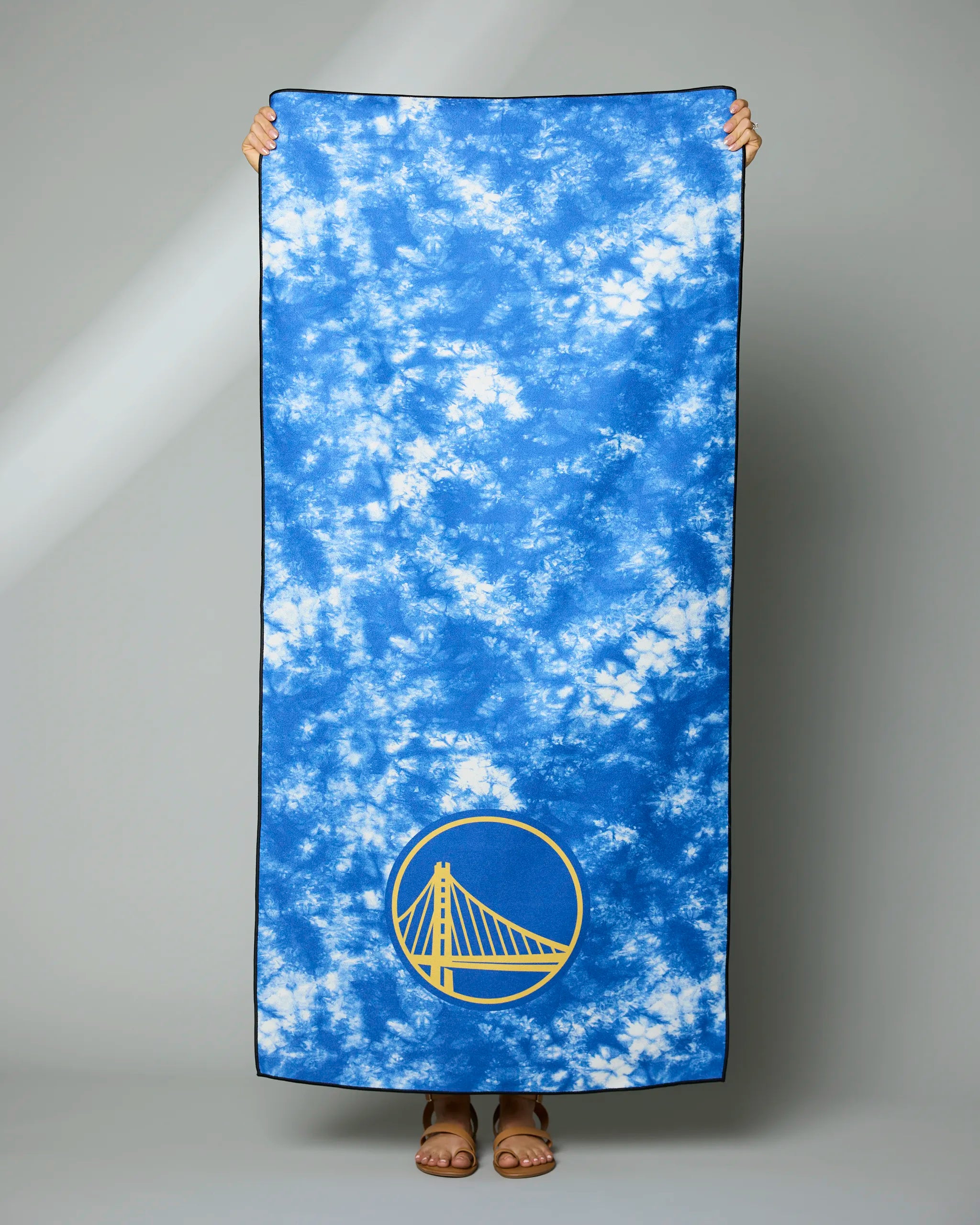 A person holds up the Geometry Warriors Flash towel—a large blue and white tie-dye design with the Golden State Warriors logo in yellow and blue at the corner. Only their hands and feet are shown.