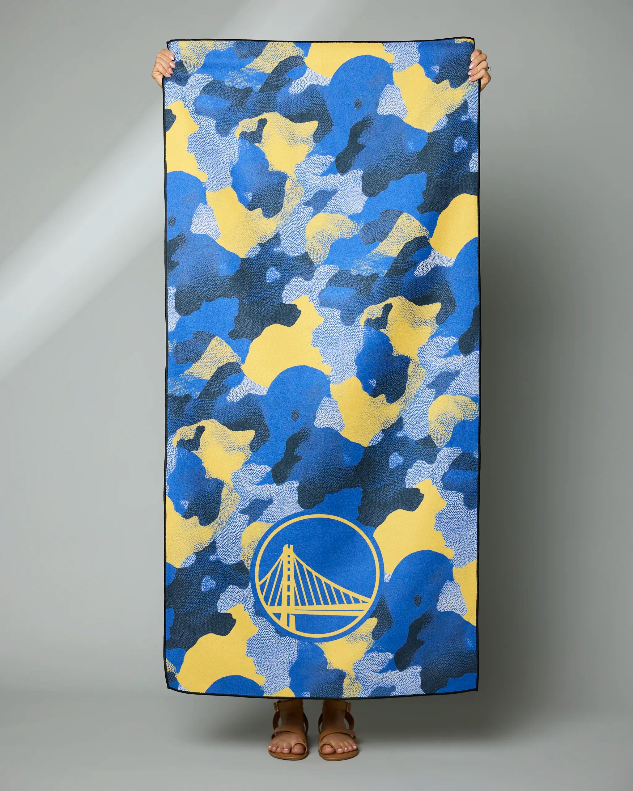 A person holds the Geometry Warriors Camo beach towel, featuring a blue, yellow, and black camo pattern with the Golden State Warriors logo near the bottom. Only their hands and feet are visible.