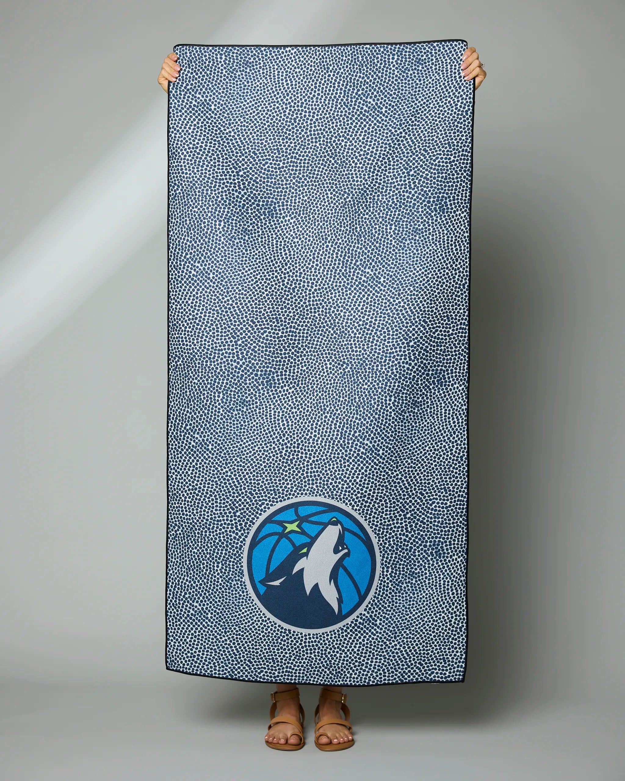 A person displays the Geometry Timberwolves Grip, a rectangular blue and white speckled blanket featuring the Minnesota Timberwolves logo near the bottom. Only their hands and sandaled feet are visible against a plain gray background.